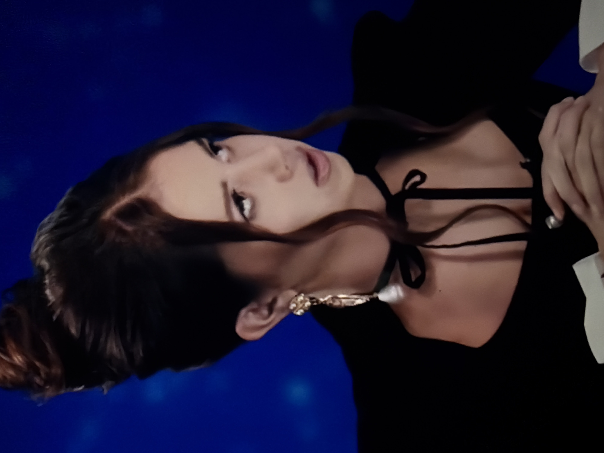 A close-up, portrait-style image features a woman with dark hair, looking upwards and slightly to her right. Her mouth is slightly open, conveying an expressive or pensive mood. She wears a black top with a plunging neckline and a black ribbon or tie detail around her neck, resembling a choker. A prominent, ornate gold earring with a white, possibly pearl-like, drop is visible on her right ear. Her dark hair is styled with volume and some strands gracefully frame her face. The background is a solid, deep royal blue, accented with subtle, diffused lighter blue specks, giving it a soft, slightly luminous quality. A portion of a hand is visible on the far right side of the frame. The location context for this image is North Kuta, Indonesia. There is no visible text in the image.
