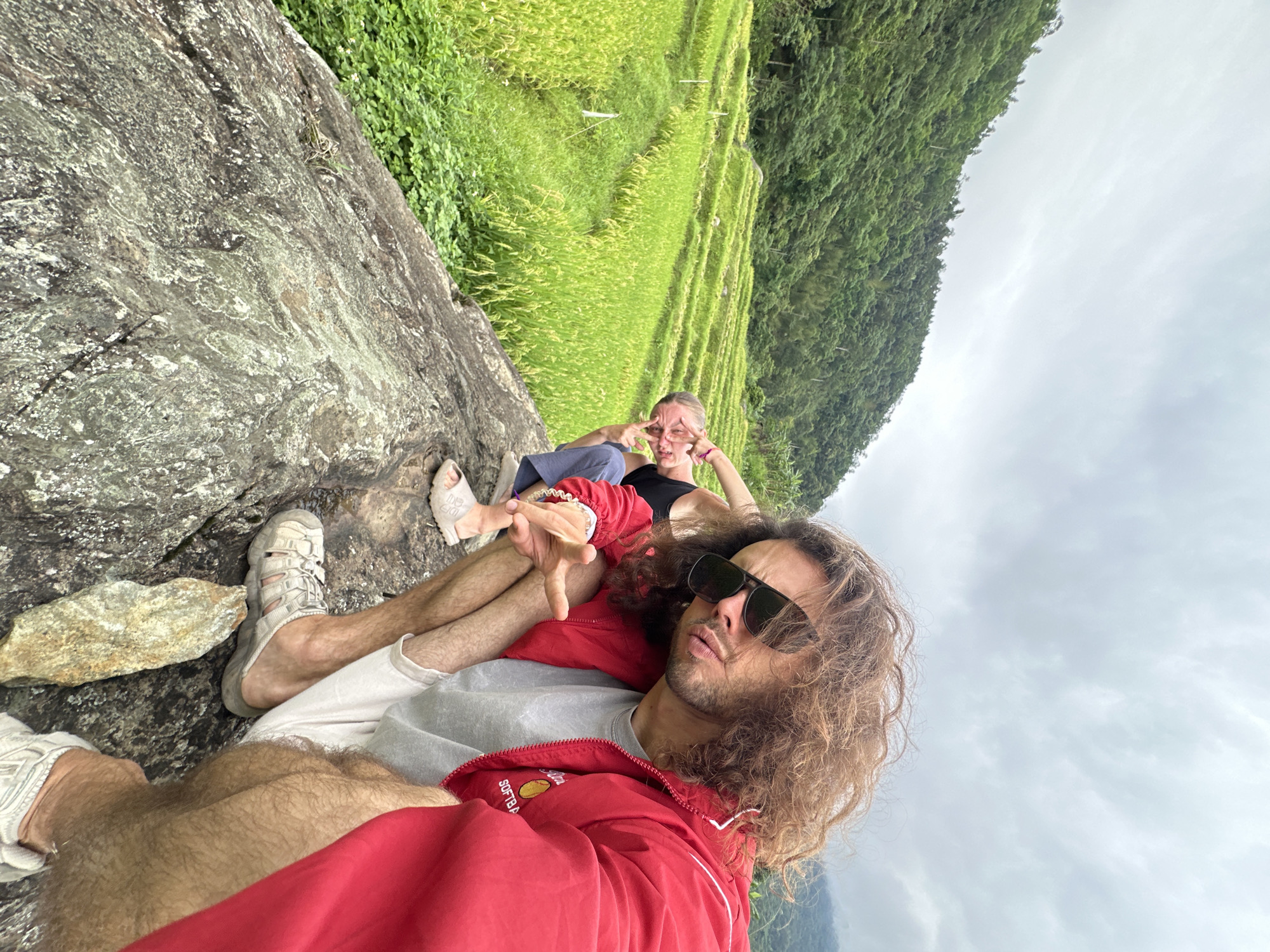 Two individuals are positioned on a large rock overlooking terraced rice fields in Pù Luông Commune, Vietnam. The person in the foreground is a male with long, curly brown hair and sunglasses, wearing a red jacket over a grey t-shirt and white shorts. He is crouching, with his left hand gesturing forward. Behind him, to the left, another individual, appearing to be a female, is also crouching. She is wearing a black top and blue pants, and her hands are near her face in a playful gesture. The rice paddies are vibrant green, arranged in distinct terraces on a hillside covered with lush vegetation. The sky is overcast, suggesting a cloudy day.