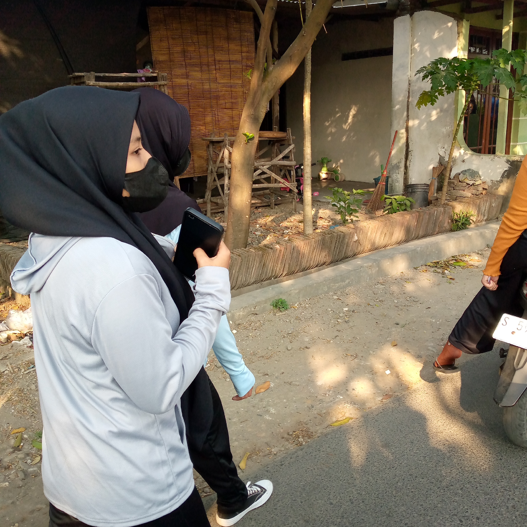 In Tambakagung, Indonesia, two individuals are captured in a side profile view, walking along a paved street. The person in the foreground, wearing a grey hoodie and black hijab, is holding a black smartphone. Beside them, another person in a dark hijab walks partially obscured. The environment suggests a residential or semi-rural area, with a weathered bamboo screen structure, a tree with a wooden support, and a house visible in the background. A parked motorcycle with a license plate that reads "S 5" is also present. The ground is a mix of asphalt and dirt, with some debris and fallen leaves scattered around. The lighting indicates it is daytime, with shadows cast by the trees on the ground.