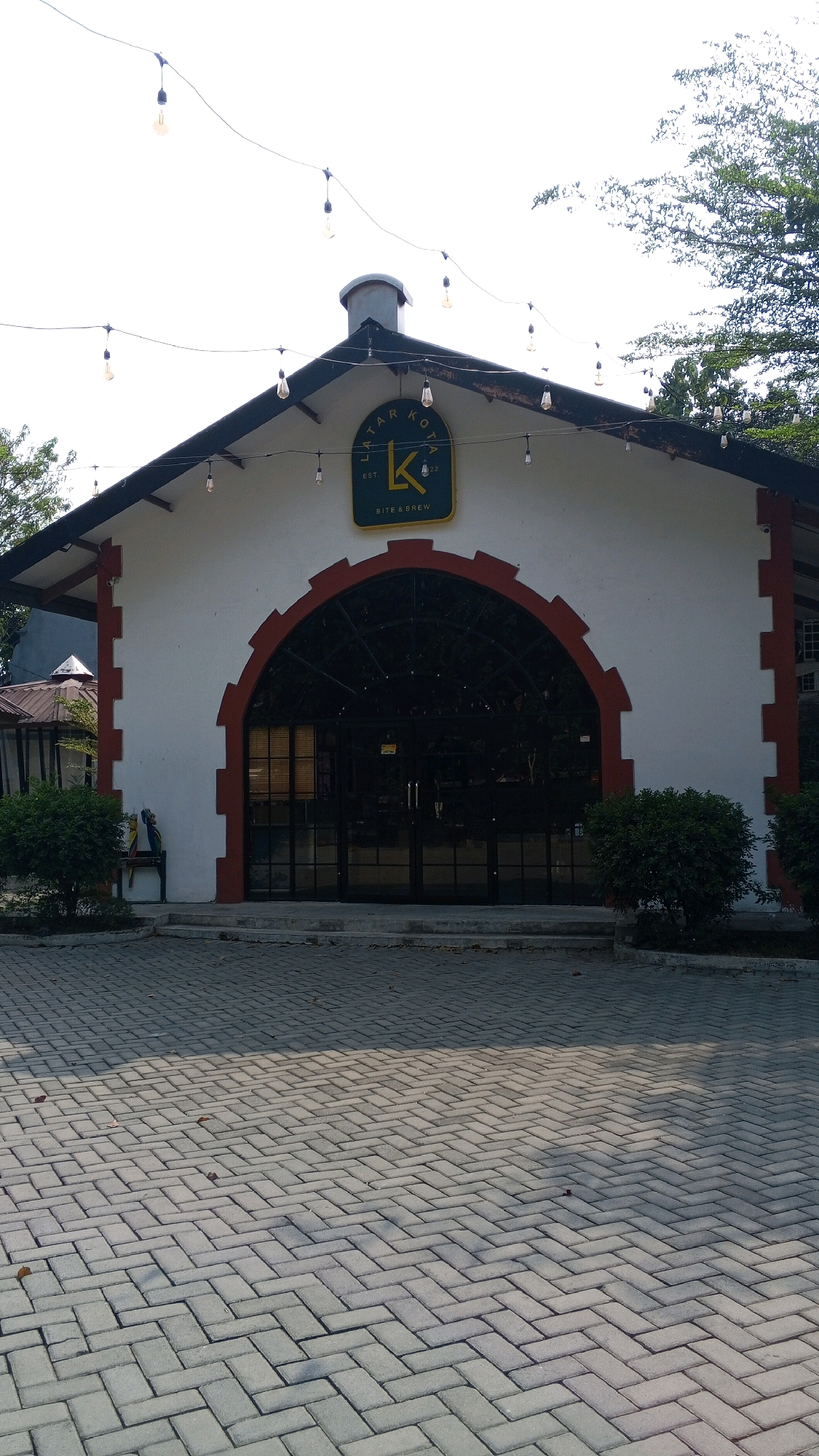 The image shows the exterior of a building with a sign that reads "LATAR KOTA BITE & BREW". The building has a white facade with red accents and a large arched entrance with glass doors. String lights are hanging above the building. The foreground is a paved area with interlocking bricks. The building appears to be a restaurant or cafe. Based on the sign "LATAR KOTA", this is likely located in Semarang, Indonesia, as "Latar Kota" translates to "City Square" or "City Background" and is a common name for public spaces and establishments in Indonesian cities. The building's architecture suggests a modern design with some rustic elements. The overall scene depicts an inviting outdoor space, possibly a popular gathering spot in the city.