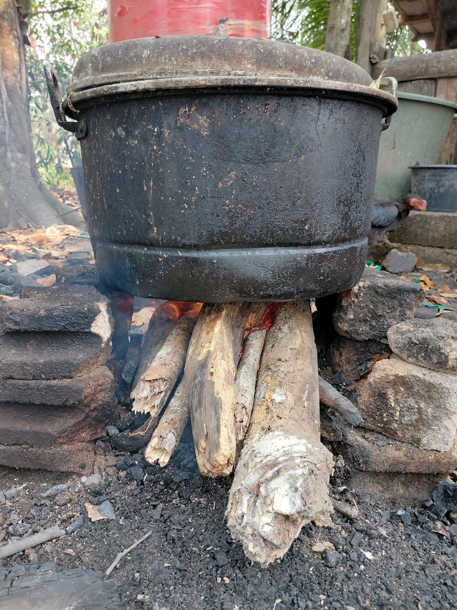 A large, black, well-used cooking pot is suspended over a fire pit constructed from rough, dark-colored stones and burning logs. The pot appears to be made of cast iron and has a weathered, smoky patina. The fire is visible beneath the pot, with flames and glowing embers illuminating the wood. The ground around the fire pit is covered with ash, charcoal, and fallen leaves. In the background, blurred foliage and trees suggest an outdoor setting. A large red cylindrical object is partially visible behind the pot, and a green plastic container and a smaller black container are also in the background. The overall scene depicts a rustic, traditional cooking setup. The location is identified as Amin Jaya, Indonesia.