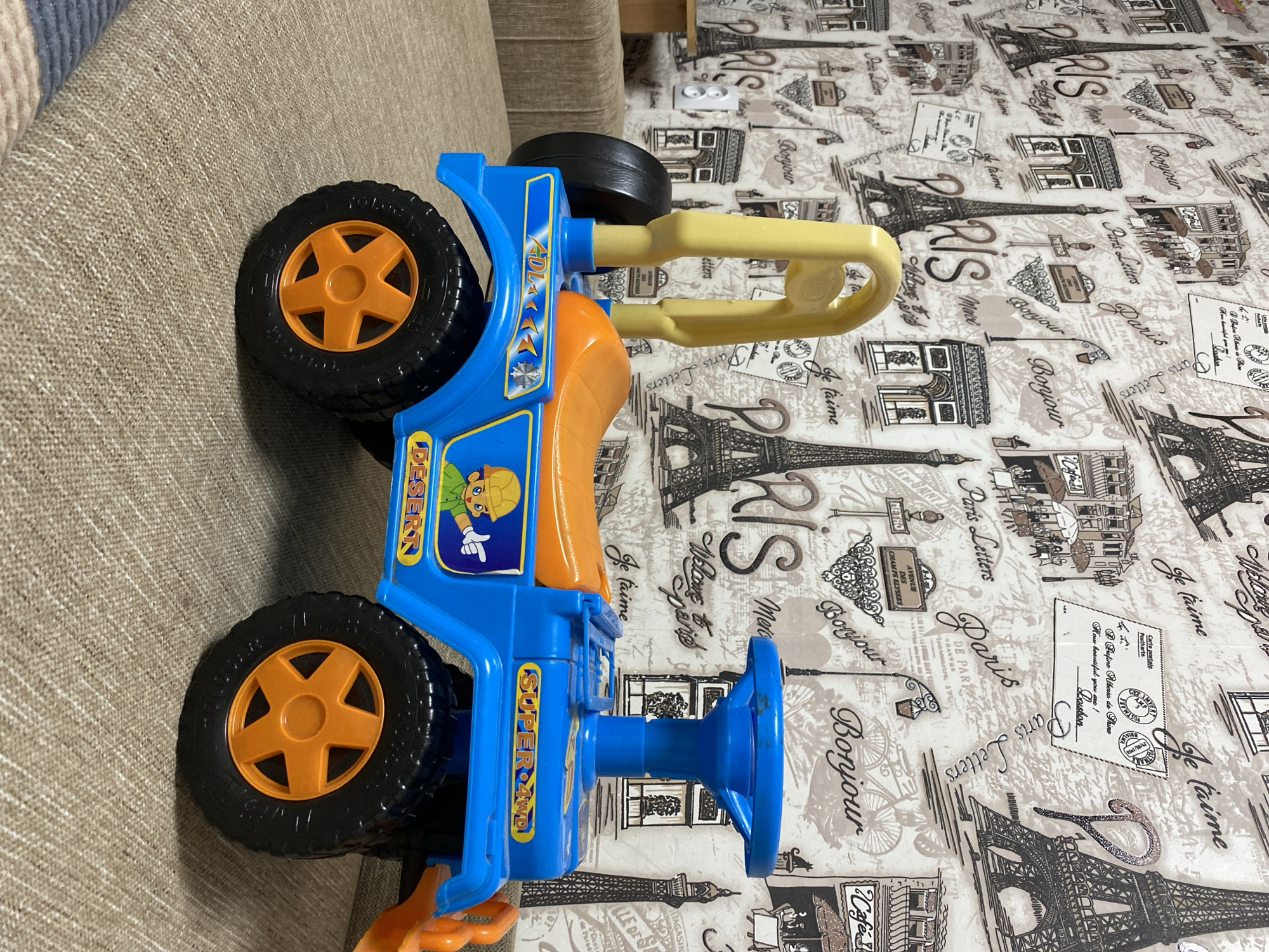 A blue toy jeep with an orange seat and steering wheel is parked on a beige, textured couch. The jeep has a yellow roll bar and orange wheels. A sticker on the side of the jeep depicts a cartoon construction worker. The background is a wallpaper with a pattern of Parisian landmarks, such as the Eiffel Tower and the Arc de Triomphe, along with French text like "Paris" and "Bonjour". There is a white electrical outlet on the wall. The scene appears to be indoors, likely in a home setting, and the location of Бескөл ауылдық округі, Kazakhstan, is not evident from the visual information provided.