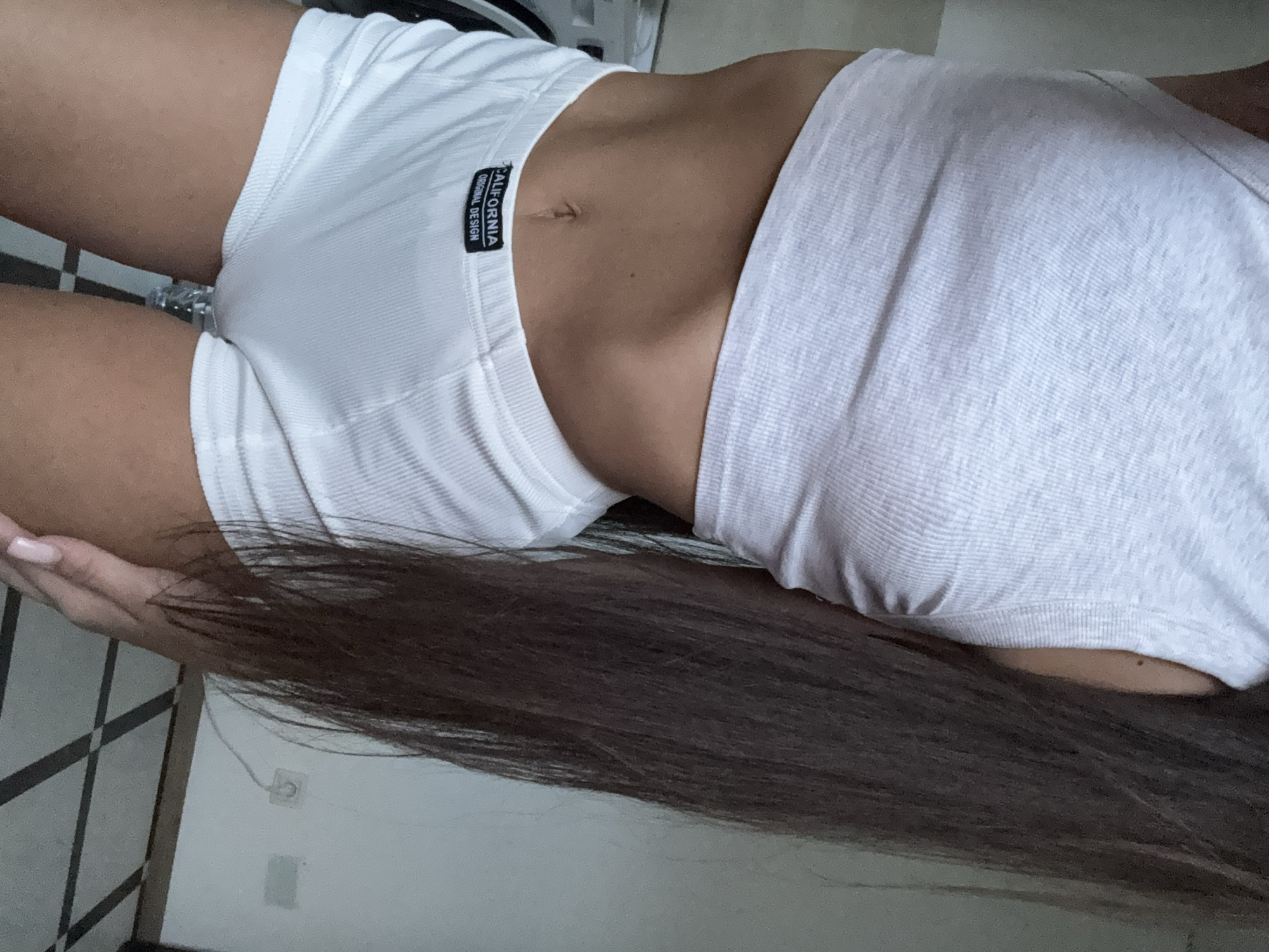 The image shows a close-up view of a person's midriff and upper thighs, wearing a white ribbed crop top and white ribbed shorts. The shorts have a black tag with white text that reads "CALIFORNIA ORIGINAL DESIGN". The person has long, dark brown hair that falls over their right shoulder. The background consists of a wall with a power outlet and a tiled floor with a black and white geometric pattern. No specific location within the city of Chișinău, Moldova can be identified from the image.