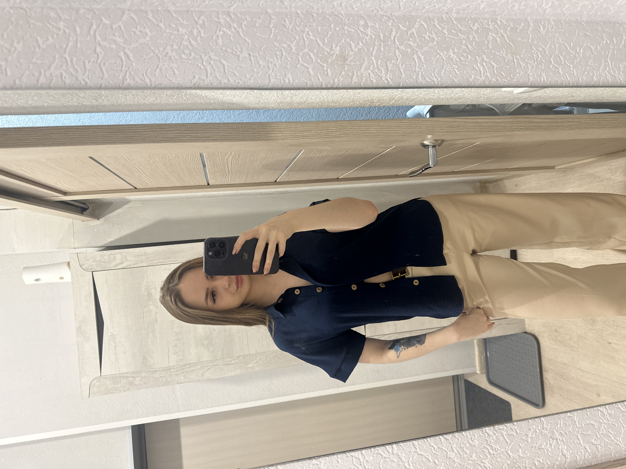 A woman in a dark blue, short-sleeved shirt and light tan pants takes a selfie in a mirror. The scene appears to be inside a bathroom in a location identified only as Unknown, Unknown. The mirror reflects a neutral-colored door and wall. The woman holds a phone up to her face, partially obscuring her features. A tattoo is visible on her left arm.
