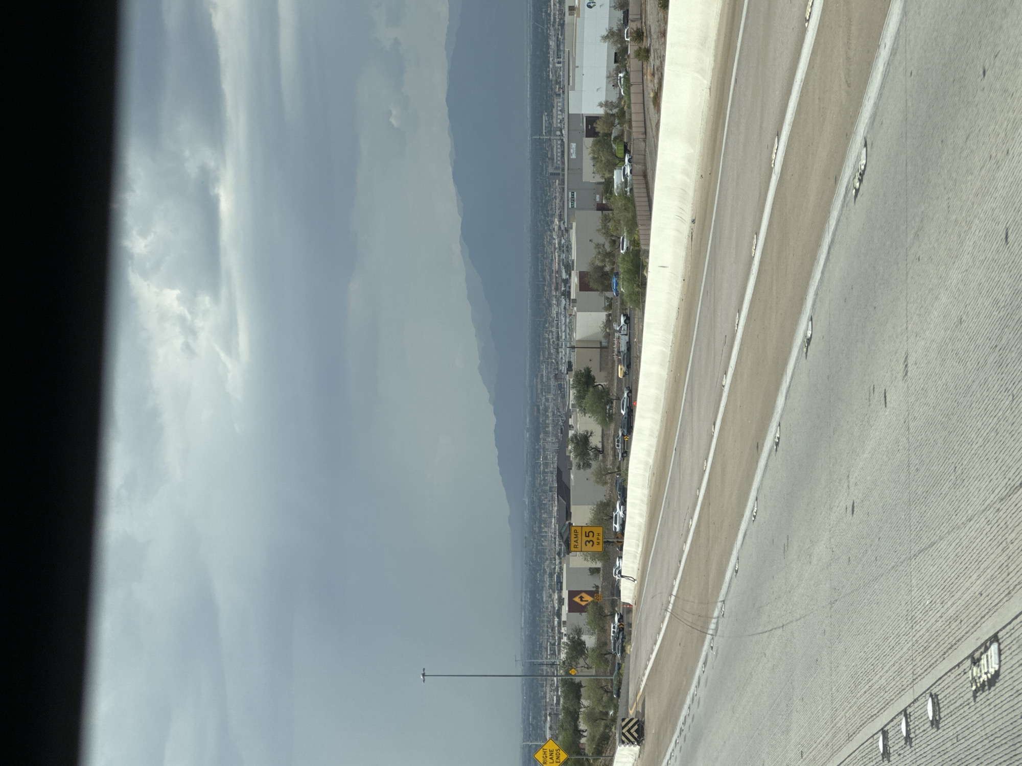 The image captures a daytime view from a vehicle on a highway in Henderson, Nevada, USA. The sky is overcast with dark, dramatic clouds that suggest an approaching storm or rain. In the foreground, the highway stretches ahead, with multiple lanes visible and reflective markers embedded in the asphalt. A concrete barrier separates the highway from the surrounding area.
Beyond the highway, a cityscape unfolds, characterized by numerous industrial or commercial buildings with light-colored facades. A line of trees and vegetation separates the buildings from the highway. In the distance, a range of mountains forms the horizon, partially obscured by haze.
Several traffic signs are visible along the side of the highway. One yellow diamond-shaped sign indicates "RIGHT LANE ENDS." Another sign displays "RAMP 35 MPH." There are also signs with directional arrows and speed limits. A line of vehicles is parked or stopped along the side of the road beyond the barrier, suggesting a potential traffic incident or a temporary stoppage. One white pickup truck is parked amongst these vehicles. The overall scene conveys a sense of urban sprawl and infrastructure against a backdrop of natural, mountainous terrain under a heavy sky.