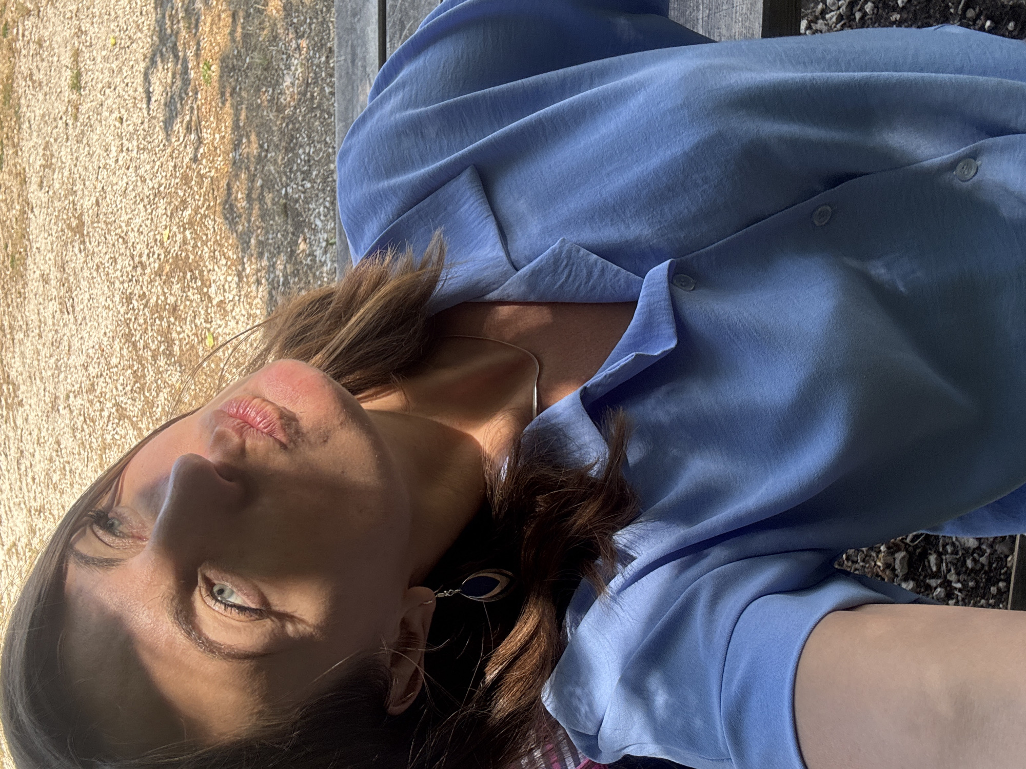 A close-up selfie shows a woman with brown hair and blue eyes, leaning her head back outdoors. She is wearing a blue collared shirt and a silver necklace. Sunlight casts a shadow across her face and highlights her features. She is outdoors, with a blurred background of gravel and greenery, suggesting a natural or park-like setting. The image was taken in Coteaux du Lizon, France.
