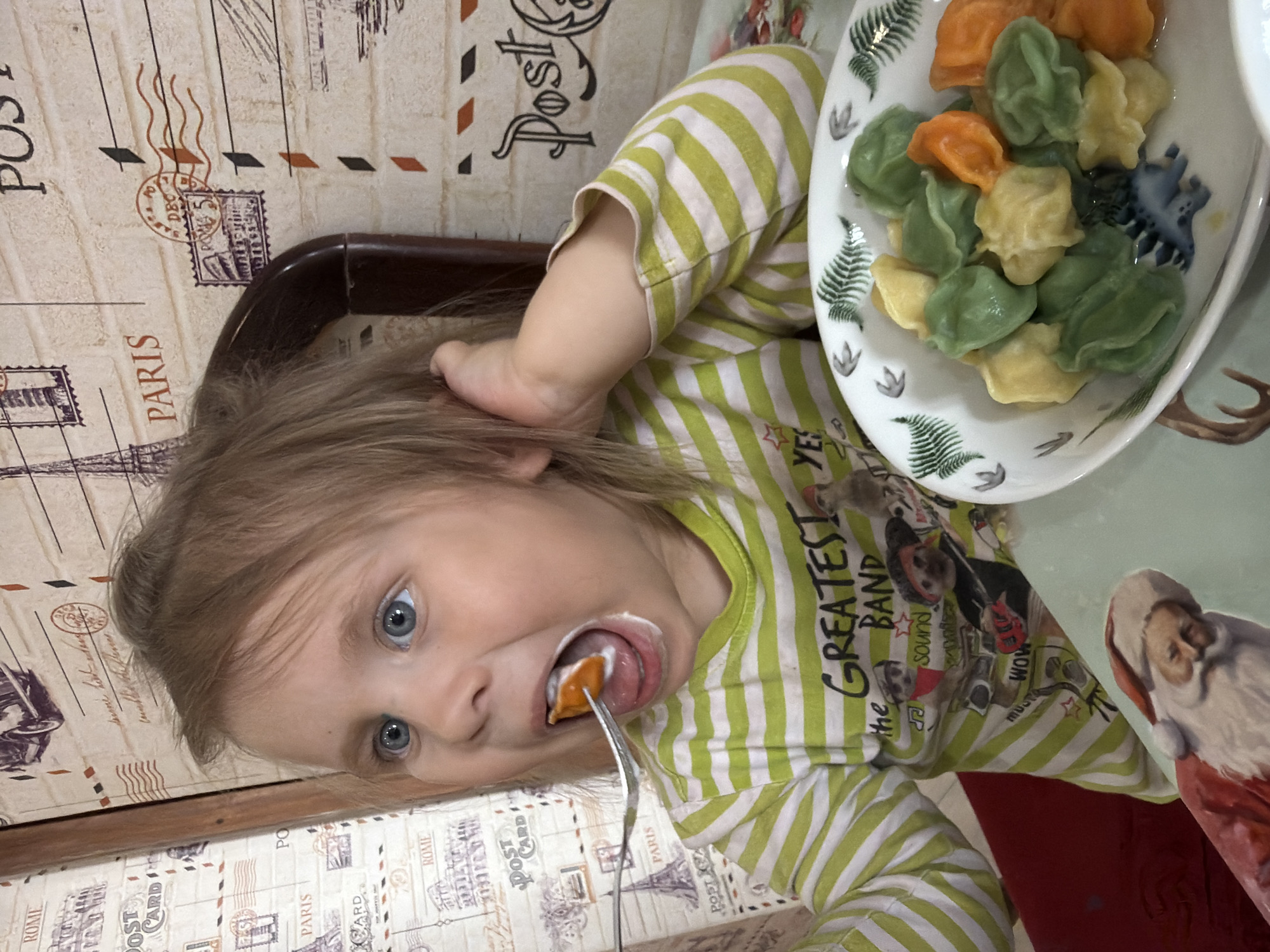 Here is a detailed description of the image:
**Scene:**
The image shows a young child eating food, seemingly at a table in a home or restaurant. The viewpoint is tilted.
**People:**
* A child with light hair and blue eyes is the central subject. They are in the act of eating with a fork.
**Subjects:**
* A plate of dumplings, colored orange, green and yellow, is present.
**Objects:**
* A fork, held by the child, with food on it.
* A plate holding the dumplings, decorated with fern and animal designs.
* A chair
* A table.
* Wall and tabletop decorated with postcard designs.
**Actions:**
* The child is eating with a fork.
**Interactions:**
* The child is interacting with their food.
**Environment:**
* The surroundings appear to be indoors. Walls and tabletop are decorated with a postcard theme. The scene is lit with overhead lighting.
**Location:**
While it is impossible to definitively confirm the location using visual information alone, the OCR text of "Paris" and "Rome" suggests a decorative motif of international cities, not necessarily that the location is actually Chaikovsky, Russia. Further information about Chaikovsky, Russia is needed to confirm the location.