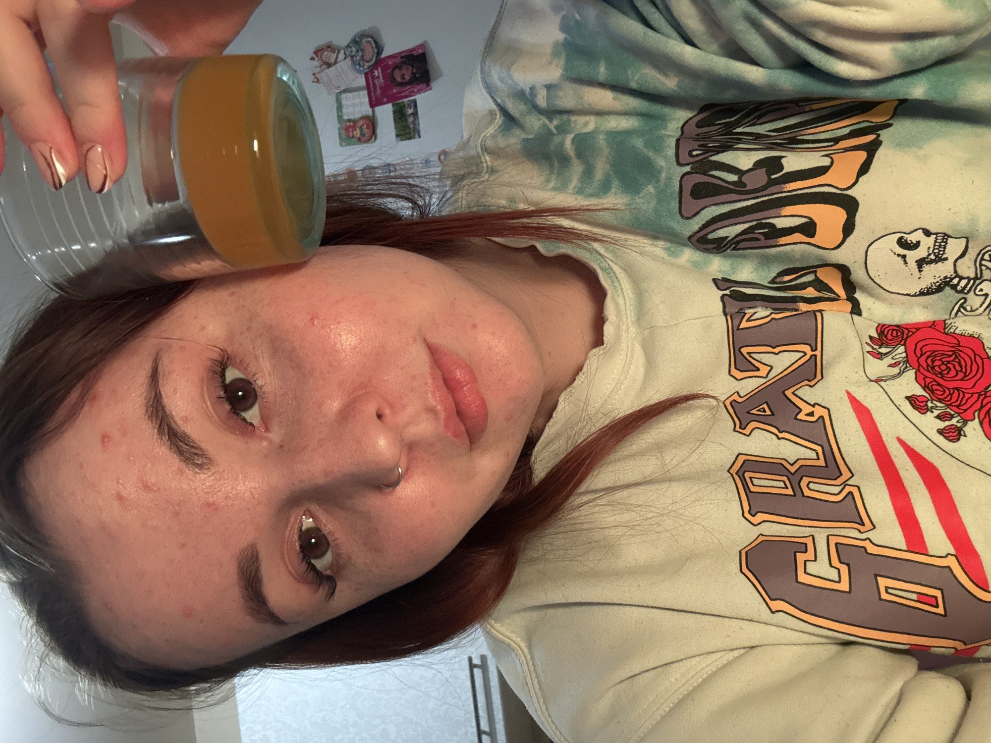 A close-up indoor selfie features a young woman with fair skin, light brown hair, and dark brown eyes, looking directly at the camera. She has a small silver hoop nose piercing on her left nostril, and minor skin blemishes are visible on her forehead and cheeks. Her lips are slightly parted and naturally full.
She holds a clear glass with a small amount of amber liquid in her right hand, positioned above her forehead. Her fingernails are manicured with a nude base and thin dark striped accents. She is wearing a light-colored, possibly tie-dye, hooded sweatshirt. The sweatshirt's graphic design includes large, stylized letters, partially visible as "GREAT," alongside an illustration of a skull and red roses.
The background shows a plain light-colored wall adorned with several small magnets or stickers, suggesting a casual home environment. The lighting is artificial, illuminating her face directly. Her expression is neutral and engaging. This scene is set in Izhevsk, Russia.