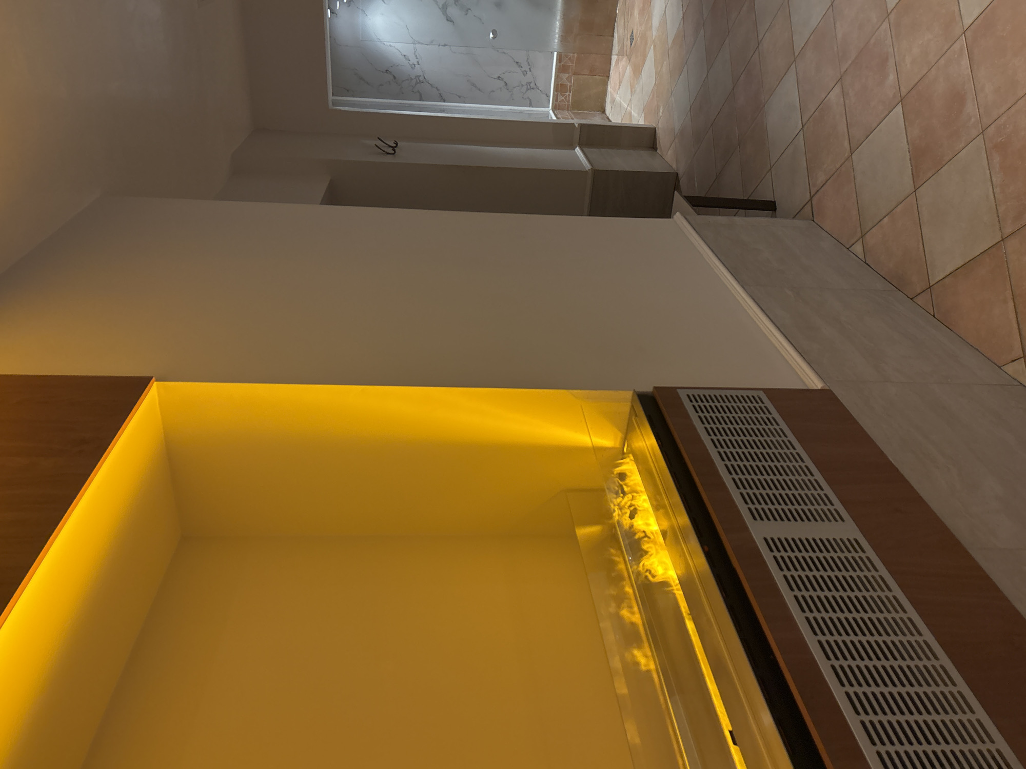 The image depicts an indoor scene, likely a spa or wellness area, with a focus on a modern fireplace with orange lighting. The fireplace is integrated into a wall unit with wooden casing and a metallic grill. The flames appear realistic, contributing to a warm ambiance. To the right of the fireplace, a tiled hallway extends into the background, leading to a shower area with marble-patterned walls and a glass enclosure. Two coat hooks are visible on a pillar in the hallway. The flooring in the hallway is made of square tiles in varying shades of beige and pinkish-brown. The walls are painted a light, neutral color. The scene is well-lit, with the warm glow of the fireplace being a prominent feature. There are no people visible in the image. This location is identified as being in Razliv, Bulgaria.