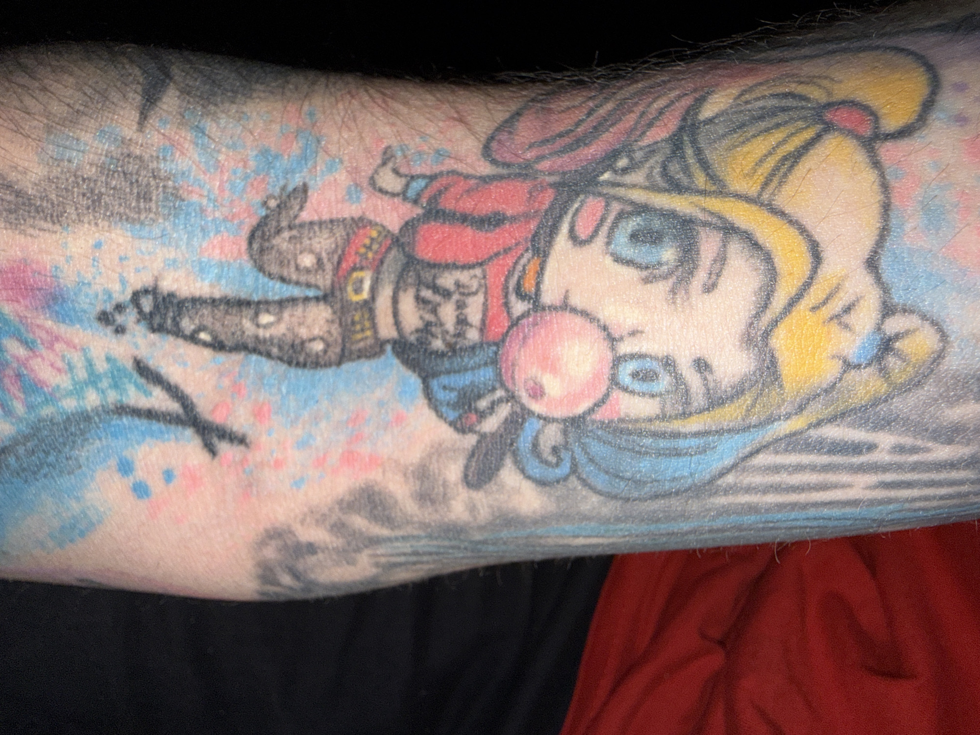 A tattoo of a cartoon character, possibly Harley Quinn, is visible on an arm. The tattoo depicts a character with blonde hair in pigtails, blue eyes, and pink lips blowing a bubblegum bubble. The character is wearing a red jacket with black accents, a black shirt with writing on it, and ripped pants with a studded belt. The tattoo has bright colors, including yellow, blue, pink, and red. The background of the tattoo appears to be abstract splashes of pink and blue. The arm is hairy and the skin tone is fair. There is a hint of a black background to the right of the arm, and a red fabric to the upper right. The location in the city of Stryama, Bulgaria, is not discernible from the provided image.