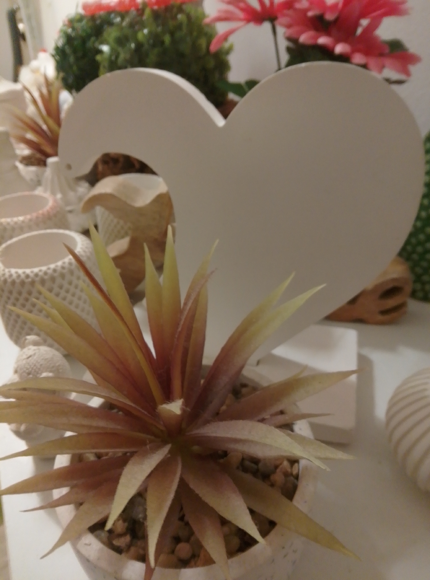 A close-up, slightly angled shot depicts a decorative arrangement on a white surface, likely a shelf or table. The central focus is a large, white, heart-shaped cutout, standing upright. In front of the heart and slightly to the left is a potted plant with spiky, thin leaves that transition from reddish-brown at the base to yellow at the tips. The pot is white and filled with small pebbles. To the left of the plant, there are several white decorative pots with textured surfaces, some appearing to be planters or candle holders. In the background, blurred pink flowers and green foliage suggest other decorative items or possibly a real plant. To the right of the heart, a wooden object with carved facial features resembling a stylized skull or mask is visible, along with a white object with a ribbed texture, perhaps a vase or candle holder. The overall impression is a collection of decorative items, possibly displayed in a shop or home. The scene is located in the city of Wedel, Germany.