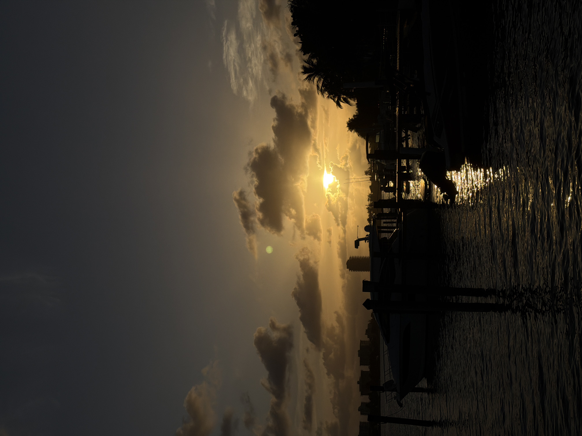 The image shows a sunset scene in what appears to be a marina or waterfront area, possibly in Miami Beach, United States. The sun is partially obscured by clouds, creating a bright glare. Silhouettes of boats, docks, and palm trees are visible along the waterfront. The water reflects the sunlight, creating a shimmering effect. In the background, the skyline of a city can be discerned through the haze of the setting sun.