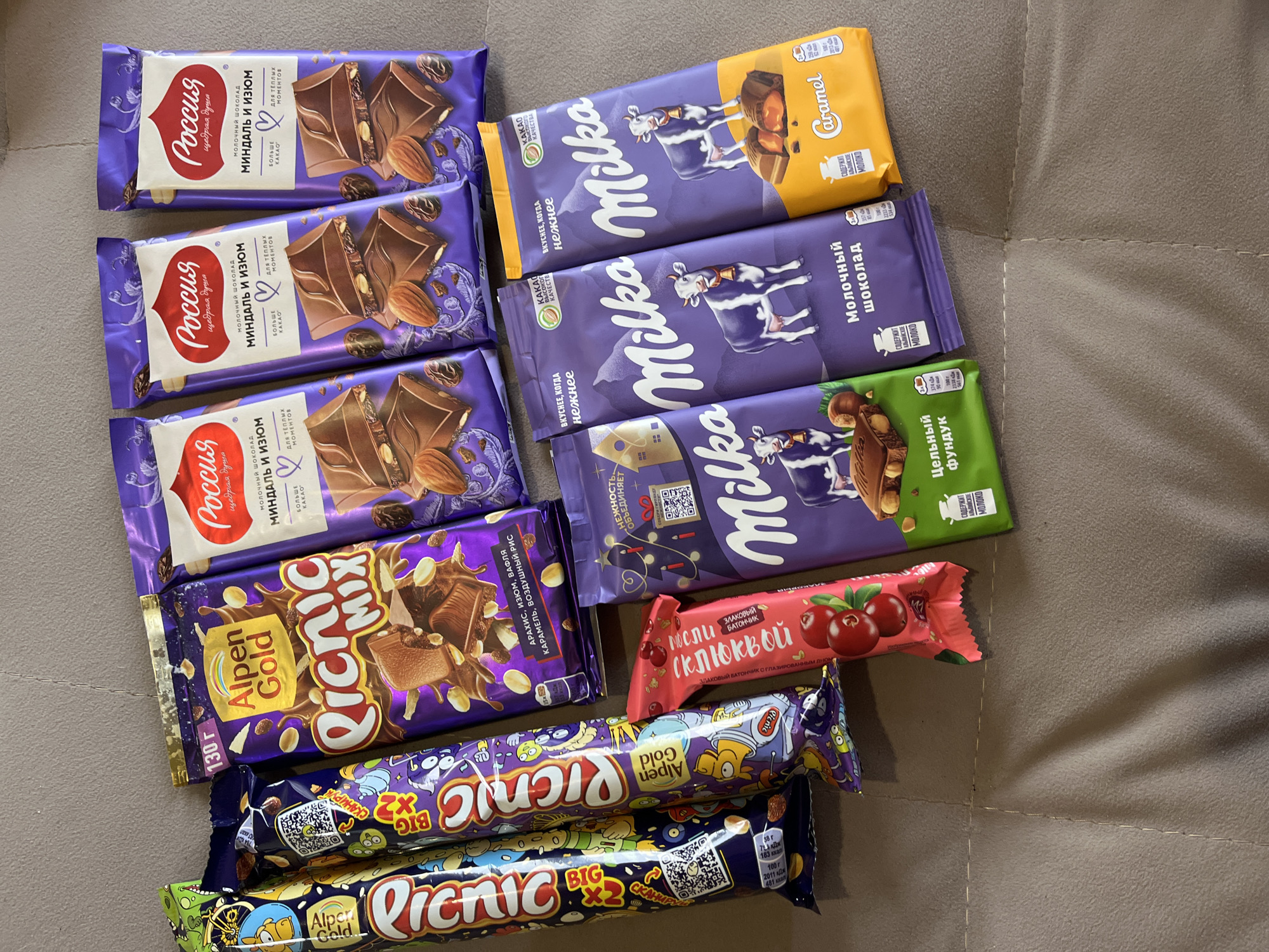 The image shows a collection of chocolate bars and snacks. Three identical chocolate bars "Россия" (Russia) with almonds and raisins are stacked on top of each other. Next to them are three Milka chocolate bars: Caramel, milk chocolate, and whole hazelnut. An Alpen Gold Picnic Mix chocolate bar is visible, along with two Alpen Gold Picnic bars. There is also a cereal bar with cranberries. The items are placed on a neutral-colored surface. The packaging is mostly in Russian. The image does not provide sufficient information to determine a location in Chaikovsky, Russia.