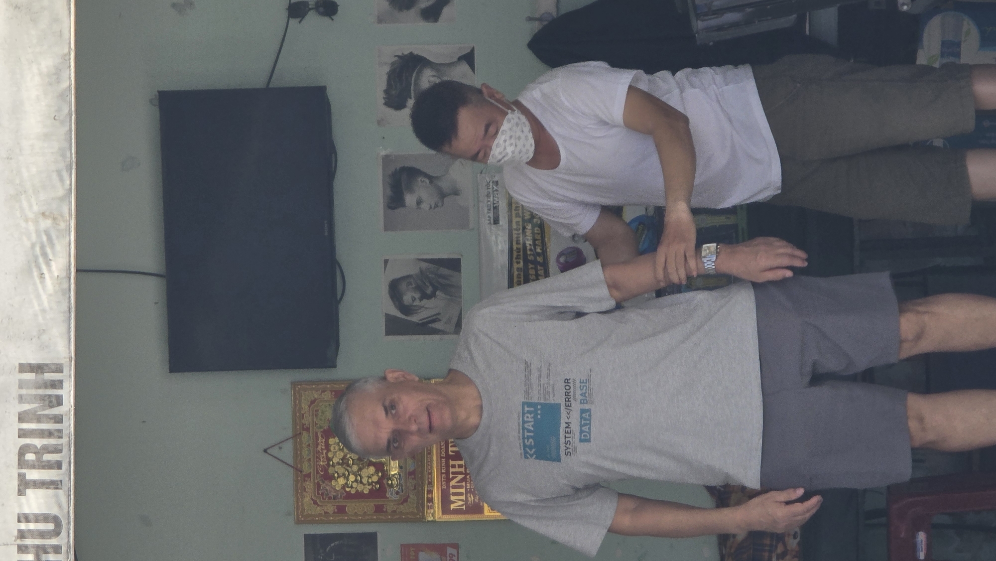 The image depicts two men in what appears to be a barber shop in Nha Trang, Vietnam. The older man is wearing a grey t-shirt and grey shorts and is standing while the younger man, wearing a white t-shirt, olive-green shorts, and a patterned face mask, is checking the older man's pulse.
The environment includes a television mounted on a light-colored wall, adorned with several framed images and posters featuring various hairstyles. There is also a sign with the words "HU TRINH" and another with the text "MINH T." The barber shop setting implies a professional interaction, with the younger man potentially attending to the older man's health or well-being.