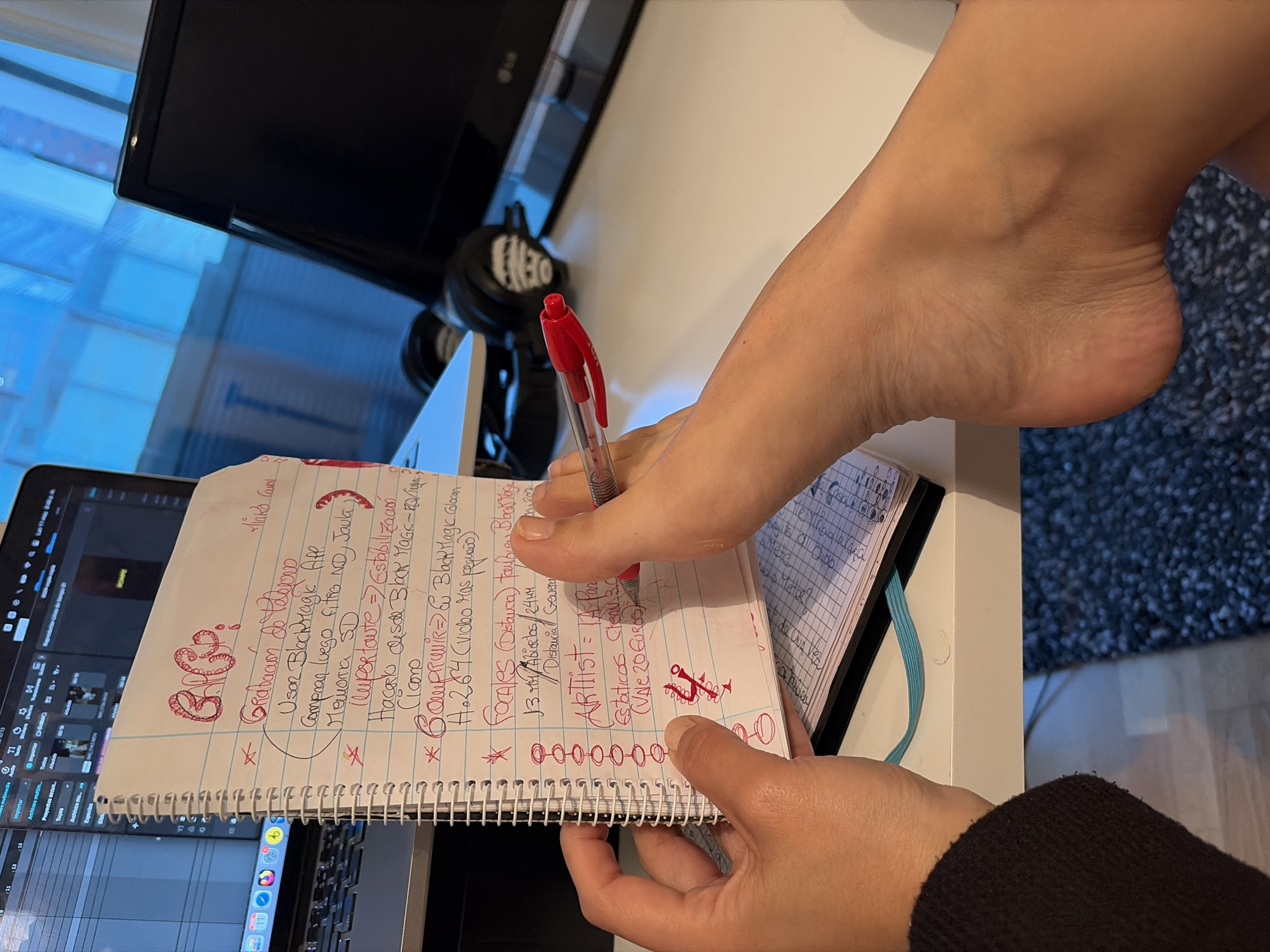 The image shows a person using a red pen to write on a notepad. The notepad is open, revealing handwritten notes in red ink, some of which appear to be in Spanish. A foot is positioned near the notepad, close to the writing hand. The notepad rests on a laptop, which displays an open video editing program. In the background, there is a computer monitor, a pair of headphones, and a window showing what looks like an urban scene with blue-tinted light. The carpet and the clothes of the person in the shot are a dark color, which creates contrast with the white desk and notebook. There is no indication that the photograph was taken in L'Hospitalet De Llobregat, Spain.