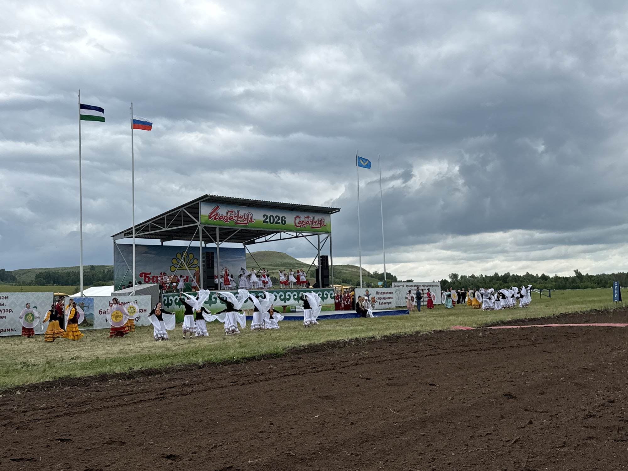 The image depicts an outdoor festival or celebration at the city of Nurkeevo, Russia, under a cloudy sky. A stage is set up on the right side of the frame, with banners that read "Нуркеево 2026" and other text in Cyrillic. Several flags are visible, including the Russian flag and another flag with horizontal stripes of blue, white, and green.
In front of the stage, a group of performers, predominantly women and girls, are engaged in a dance. They are dressed in traditional colorful attire, with some wearing white dresses and veils, while others are in vibrant yellow and red garments. They are arranged in lines and circles, performing synchronized movements.
To the left of the stage, more performers in traditional clothing stand, some holding circular props with patterns. Spectators are visible in the background, near the stage and along the perimeter of the performance area.
The environment consists of a grassy field with a strip of freshly tilled brown earth in the foreground, suggesting a rural or park-like setting. Trees and rolling hills form the backdrop behind the stage, with a few scattered structures visible in the distance. The overall atmosphere appears festive and communal.