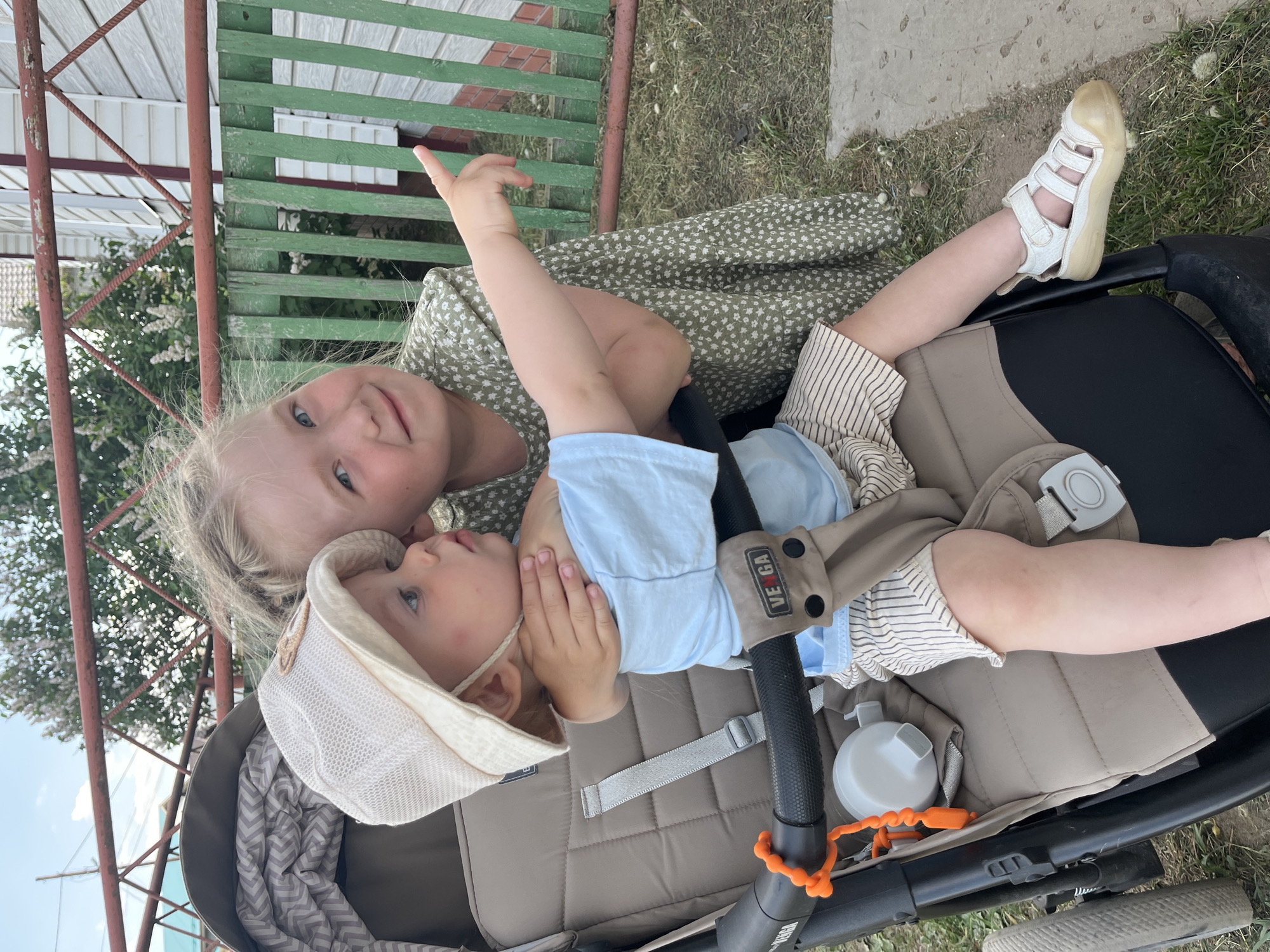The image shows two young children outdoors. A girl with blonde hair is leaning over a baby in a stroller. The baby is wearing a light blue shirt and a hat. The stroller is tan and black with a bottle attached to the handlebar. In the background is a green and brown fence and a glimpse of a building. The setting appears to be in the city of Chaikovsky, Russia.