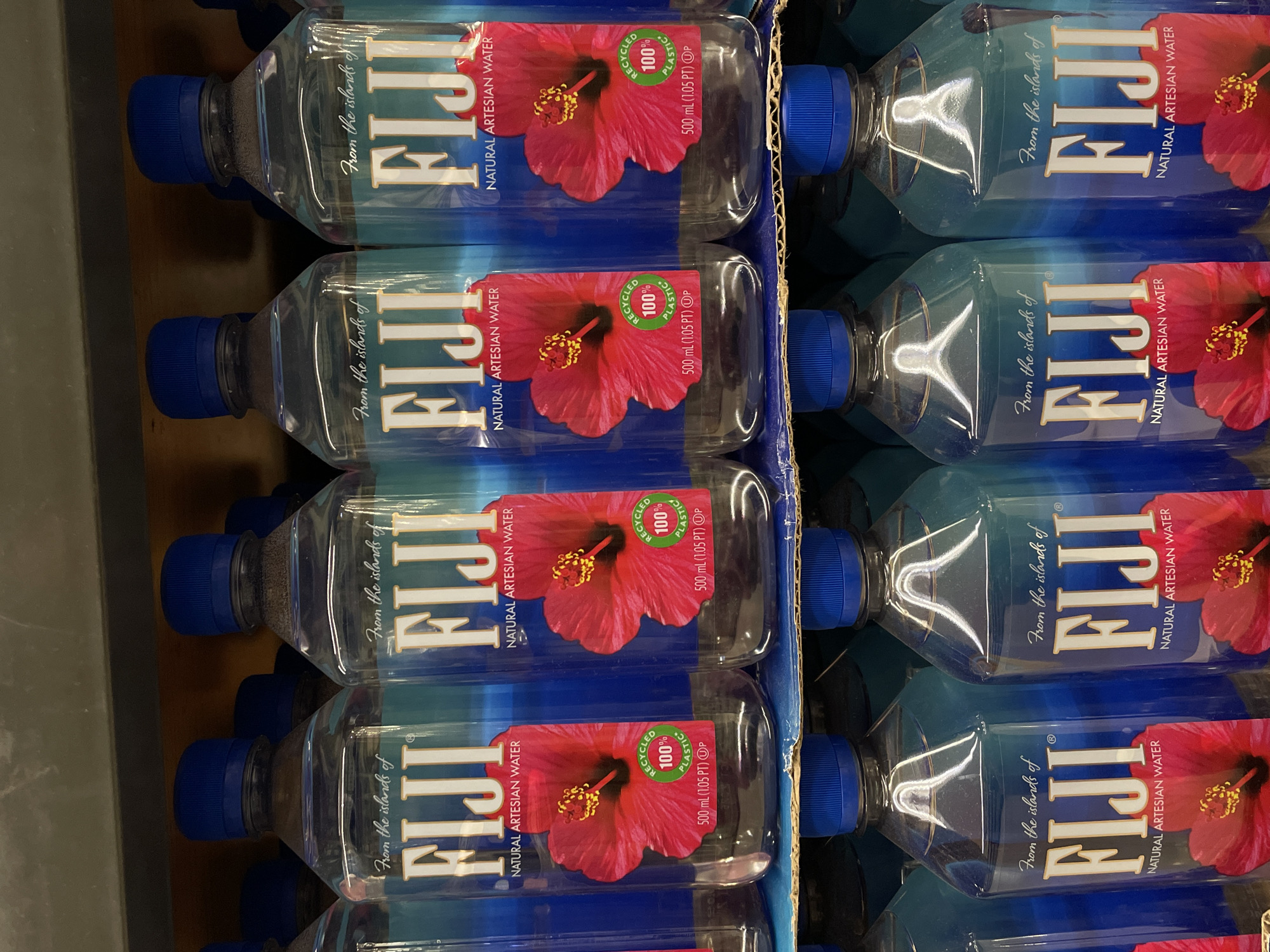 The image displays a close-up, eye-level view of several bottles of Fiji brand natural artesian water arranged on shelves. The bottles are clear plastic with blue caps and feature the distinctive Fiji branding, which includes the text "From the islands of FIJI NATURAL ARTESIAN WATER" and a red hibiscus flower graphic. Some bottles also have a circular green sticker indicating "RECYCLED 100% PLASTIC". The bottles are stacked in multiple rows, suggesting they are in a retail environment like a supermarket or convenience store. The lighting suggests an indoor setting, possibly a grocery store aisle with artificial lighting. The specific location in Davao City, Philippines is not identifiable from the visual information provided in the image.