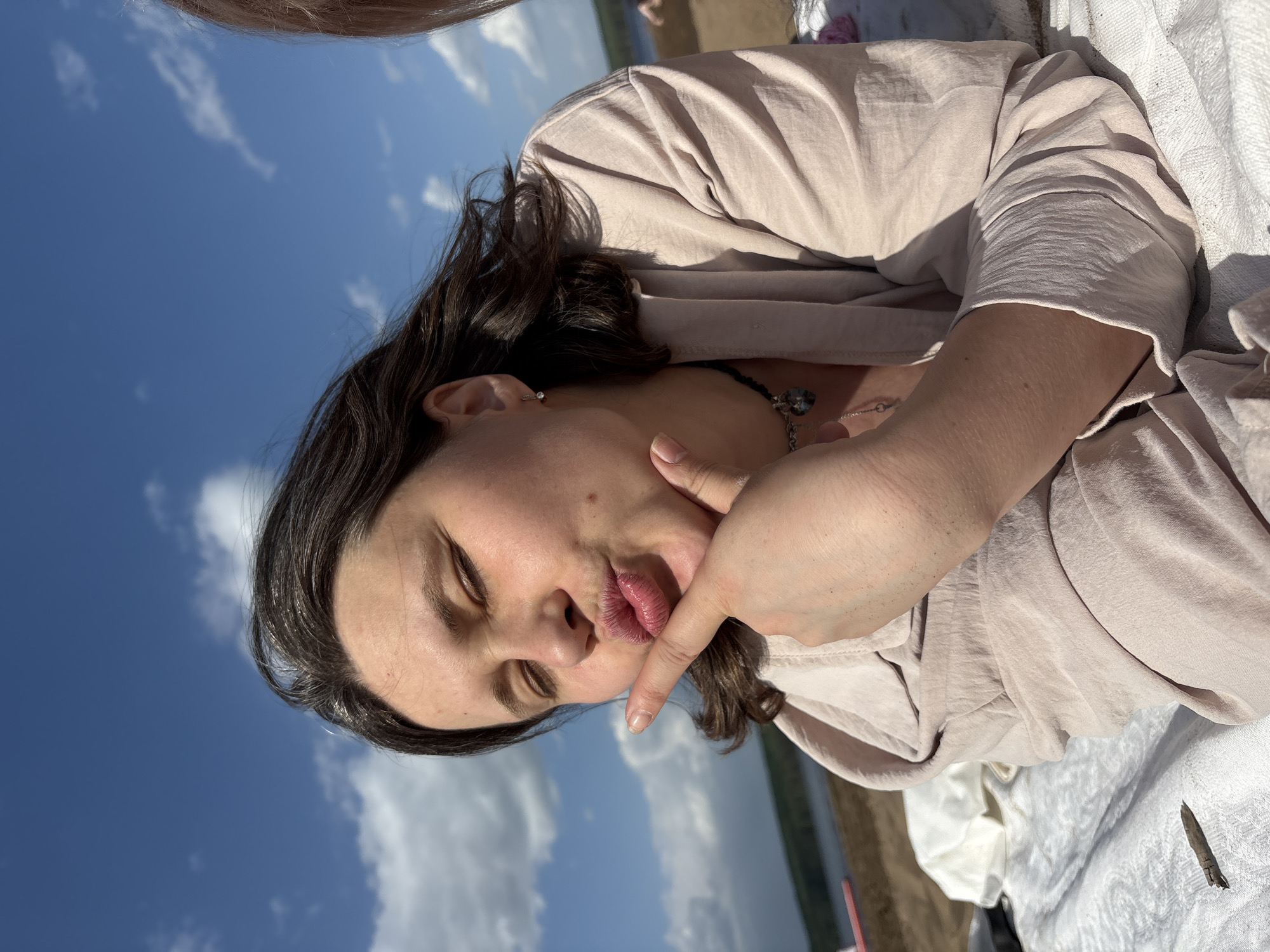 A person with dark hair and wearing a light pink shirt is lying down. Their eyes are closed and their lips are pursed. One hand is near their mouth. The background shows a blue sky with white clouds, suggesting an outdoor location, possibly a beach or park. A body of water is partially visible in the distance. The photo was taken in the city of Chaikovsky, Russia.