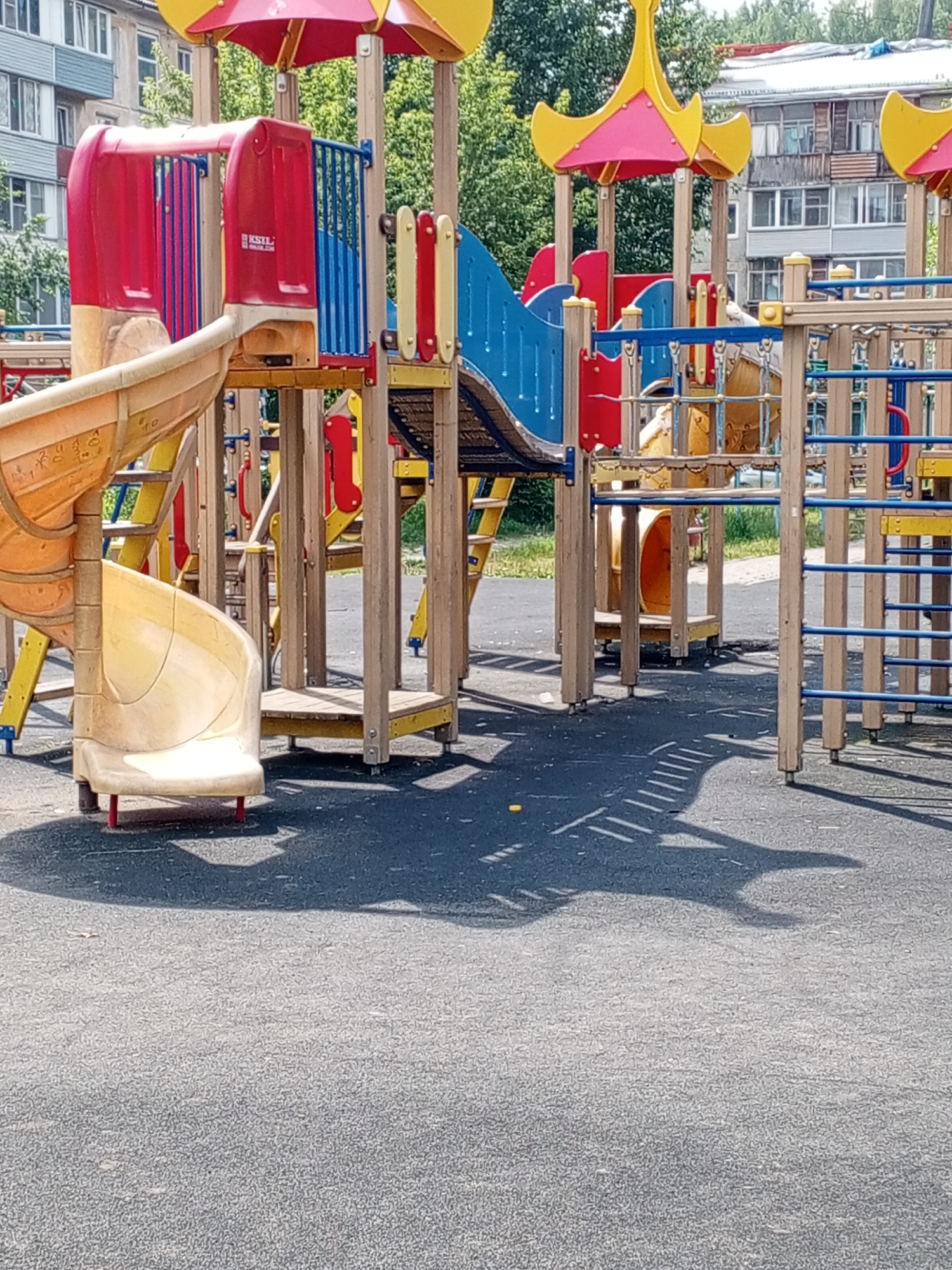 The image depicts a modern, colorful children's playground structure located in the city of Krasnoyarsk, Russia. The playground features a multi-level design with various elements such as slides, climbing structures, and safety railings. The primary colors of the playground equipment are red, yellow, and blue, set against a backdrop of green trees and residential buildings. The ground surface is a dark, textured material, possibly rubberized safety surfacing common in playgrounds. Shadows cast by the playground equipment are visible on the ground, indicating a sunny day. No people are present in the image. The residential buildings in the background appear to be typical of urban Russian architecture.