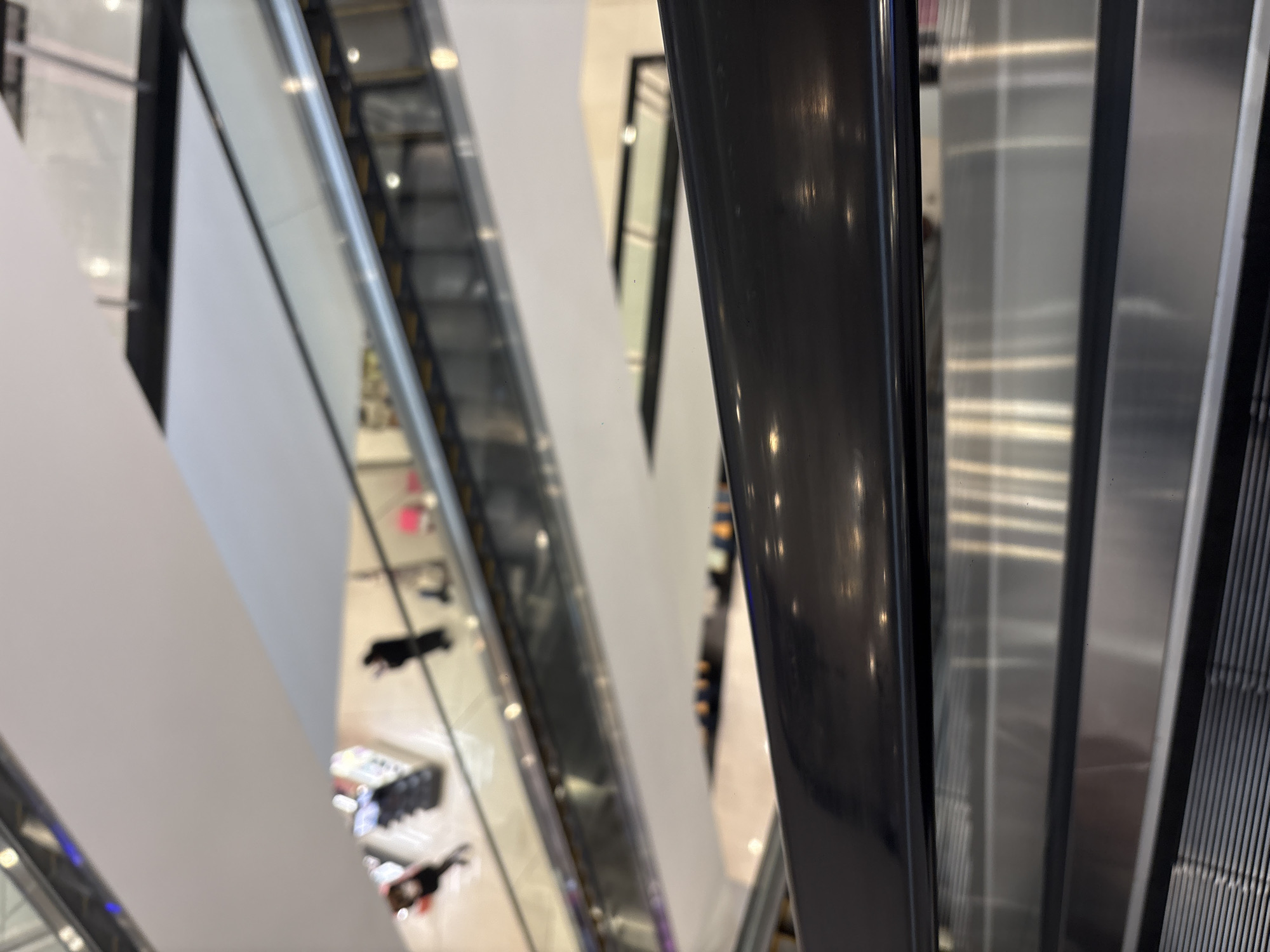 The image shows a view down several floors from the top of an escalator in a shopping center. Escalators are visible in the foreground and background, with some in motion. The shopping center contains retail establishments selling clothing and accessories. The location is most likely in Mexico City, Mexico.