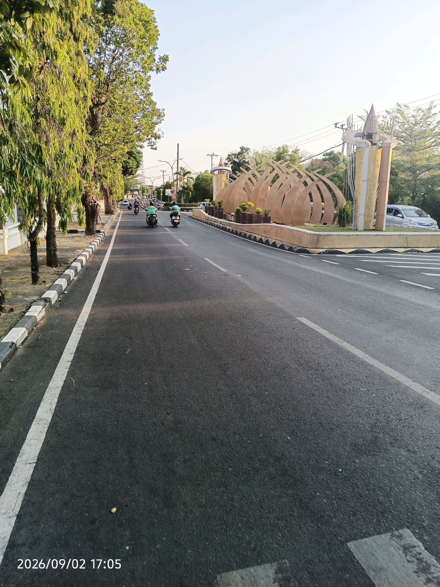 A paved road curves to the right under a clear sky during daylight. On the left, a row of tall trees with lush green foliage lines the sidewalk, which is bordered by a curb with black and white stripes. Beyond the sidewalk, a white building is partially visible. Several motorcycles are traveling on the road, with two riders in the foreground wearing green shirts. On the right side of the road, a modern decorative monument with curved, golden-brown elements and vertical supports stands. A silver car is parked near the monument. The image is dated "2026/09/02 17:05". This scene is located in Pare-Pare, Indonesia, specifically at the Tugu Apreseasi Km 5, a landmark entrance to the city.