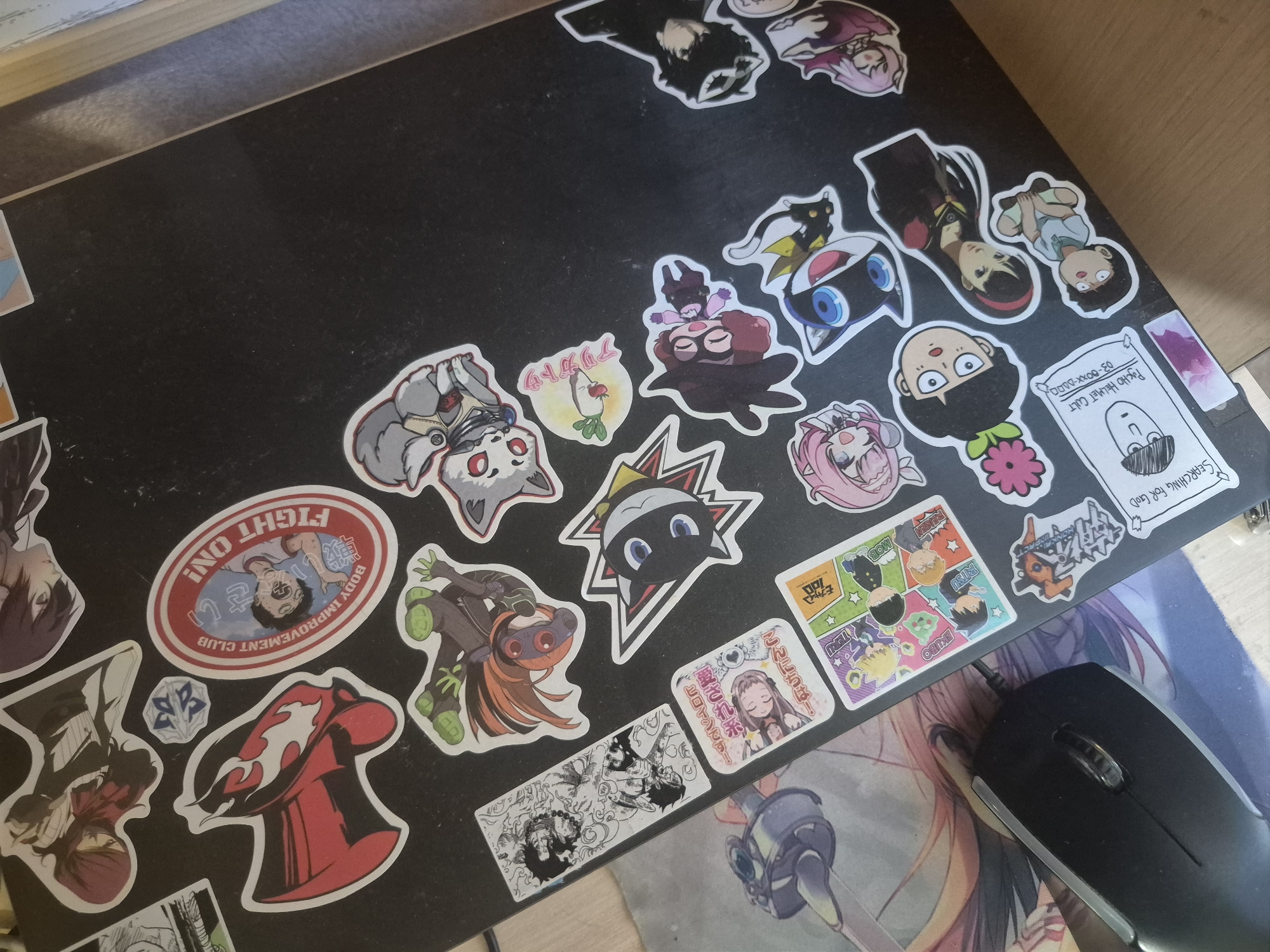 The image depicts a black surface covered in a variety of anime-themed stickers. These stickers include characters from various anime and manga series. Prominent among them are stickers featuring characters and logos from "Mob Psycho 100" (e.g., Ekubo, Ritsu, the Psycho Helmet Cult logo) and "Persona 5" (e.g., Morgana, character masks). There are also stickers with images and text, such as "FIGHT ON! BODY IMPROVEMENT CLUB" and a sticker featuring a girl saying "Hello! I'm the heroine! Thank you!" A computer mouse sits on a mousepad with an anime-style graphic, visible in the lower-right portion of the image. The arrangement suggests the surface is likely a laptop lid or a similar area where someone has applied the stickers as decoration.