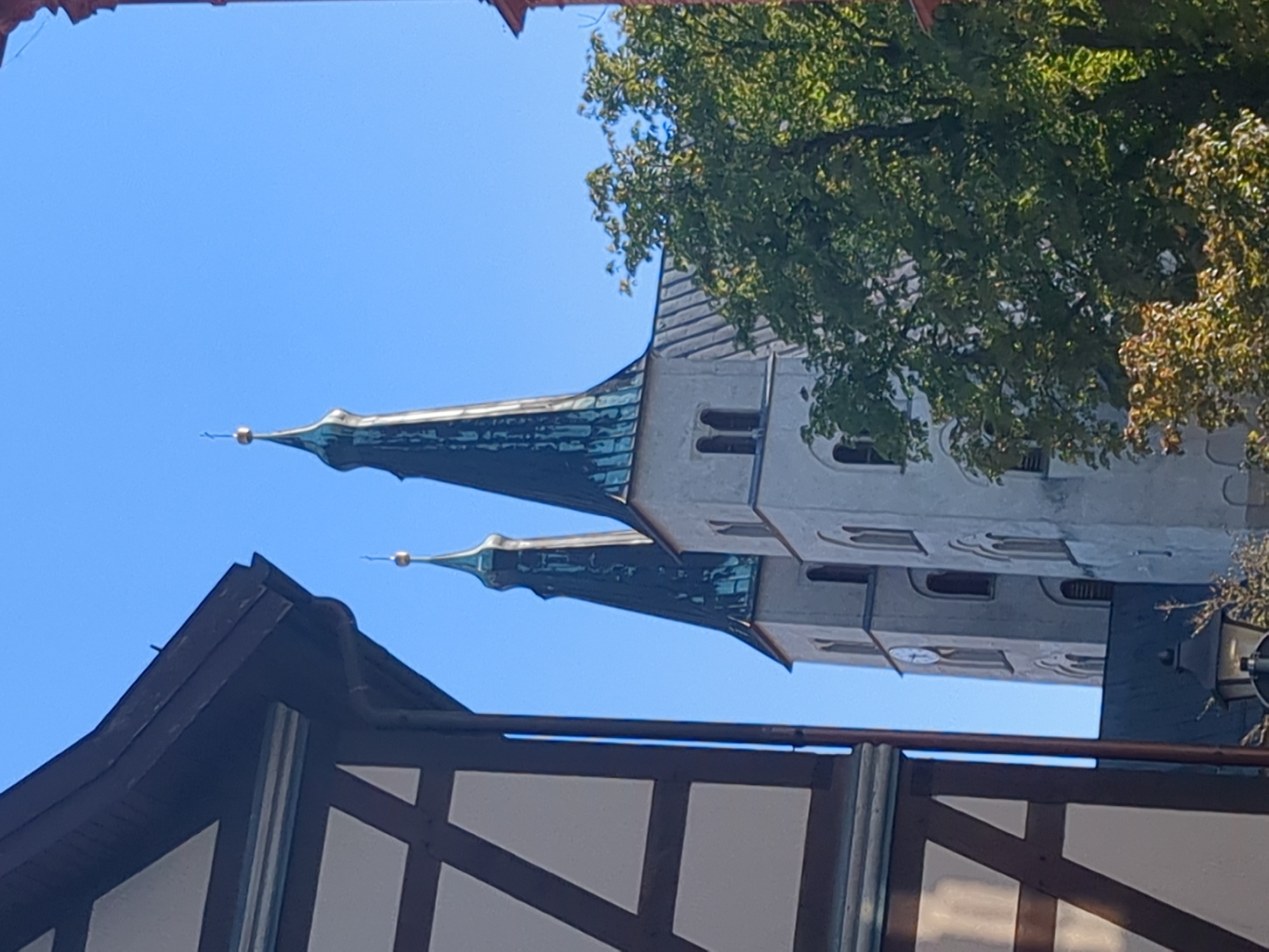 The image shows two church spires in Nordhausen, Germany, partially obscured by the corner of a half-timbered building in the foreground and a tree on the right. The spires are tall and slender, tapering to points topped with golden crosses. They are dark green and have a weathered appearance, suggesting they are made of copper or a similar material that has oxidized. The main body of the church appears to be made of light-colored stone, with arched windows and a clock face visible on the left spire. The sky is clear and blue. The half-timbered building has a dark brown wooden frame and white infill. The tree has lush green leaves, indicating it is likely summer or early autumn. The overall impression is one of a historic European city.