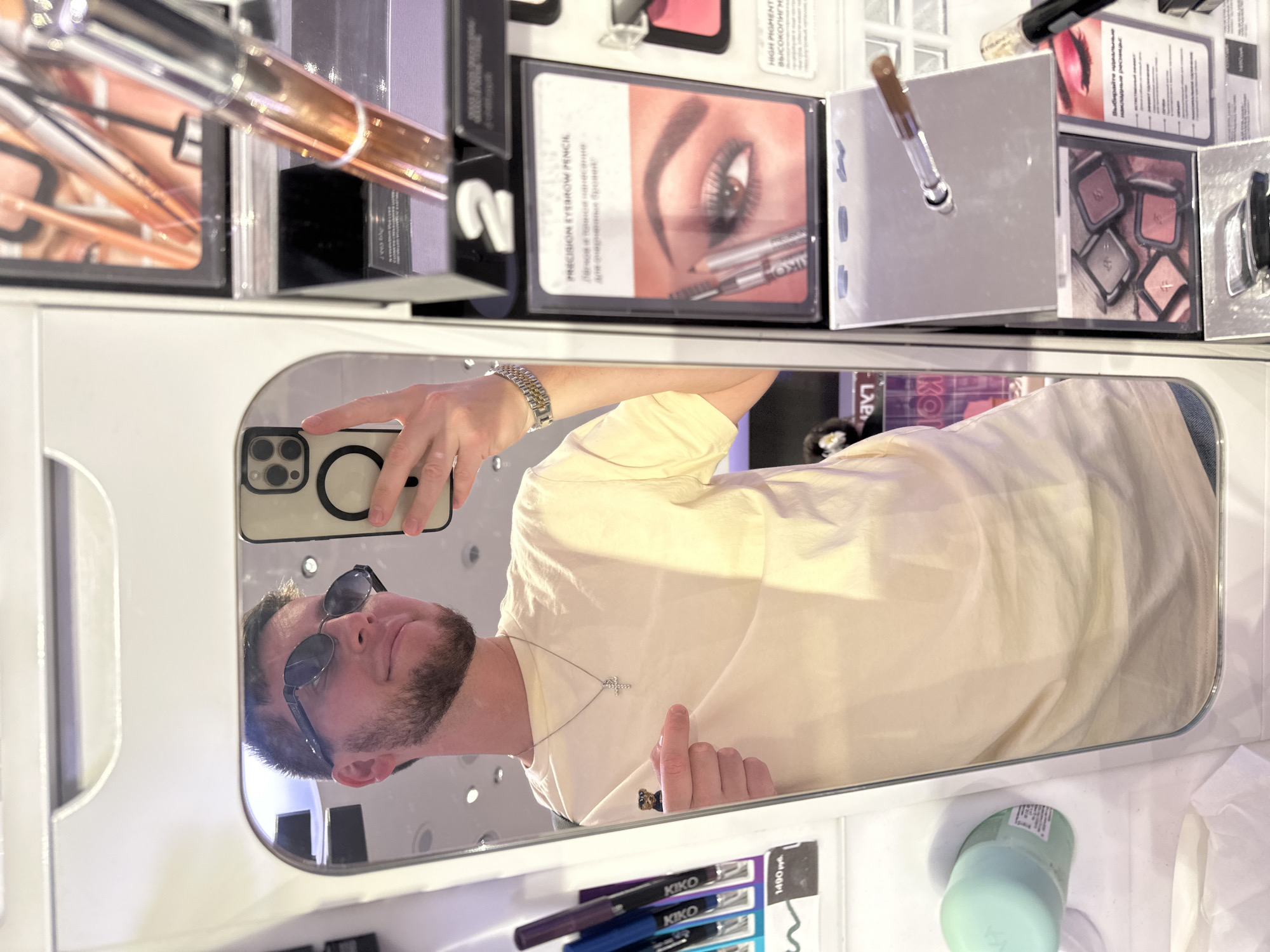 A man, with short brown hair and a beard, wearing sunglasses, a light yellow t-shirt, and a silver necklace with a cross pendant, takes a selfie in a mirror. He is wearing a silver watch on his left wrist and has a small pin on his shirt. He is pointing to his chest with his right index finger. The man is in a store, reflected in a large mirror that is part of a display. The display features various cosmetic products, including makeup brushes, eyeshadows, and eyebrow pencils, with product information visible on the packaging. In the foreground of the reflection, there are also some cosmetic products on shelves. The lighting appears to be artificial, typical of a retail environment.