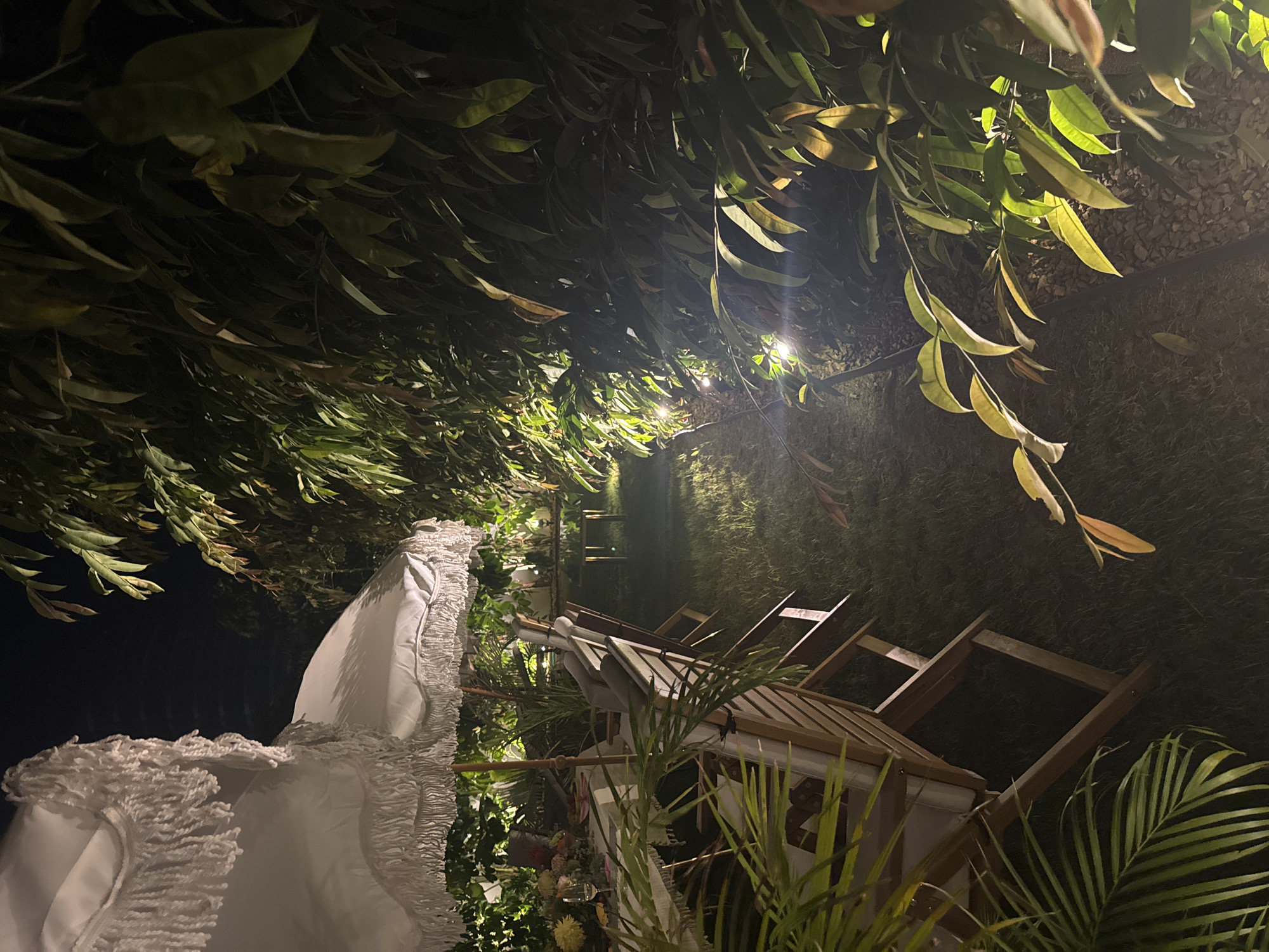 In the city of Agios Tychona Community, Cyprus, at night, a dimly lit backyard is set up for a meal. Two white umbrellas with fringed edges are partially visible, providing shade over a dining table laden with a white tablecloth, plates, glassware, and a floral arrangement. Wooden chairs with grey cushions are positioned around the table. The scene is illuminated by subtle outdoor lighting, casting a warm glow on the lush green grass and the surrounding foliage. To the right, dense green leaves form a natural wall, interspersed with small, bright lights along the base. In the background, a reflective surface, possibly a pond or pool, is visible with a small table and chair placed near its edge. The overall ambiance suggests a peaceful evening gathering in a garden setting.