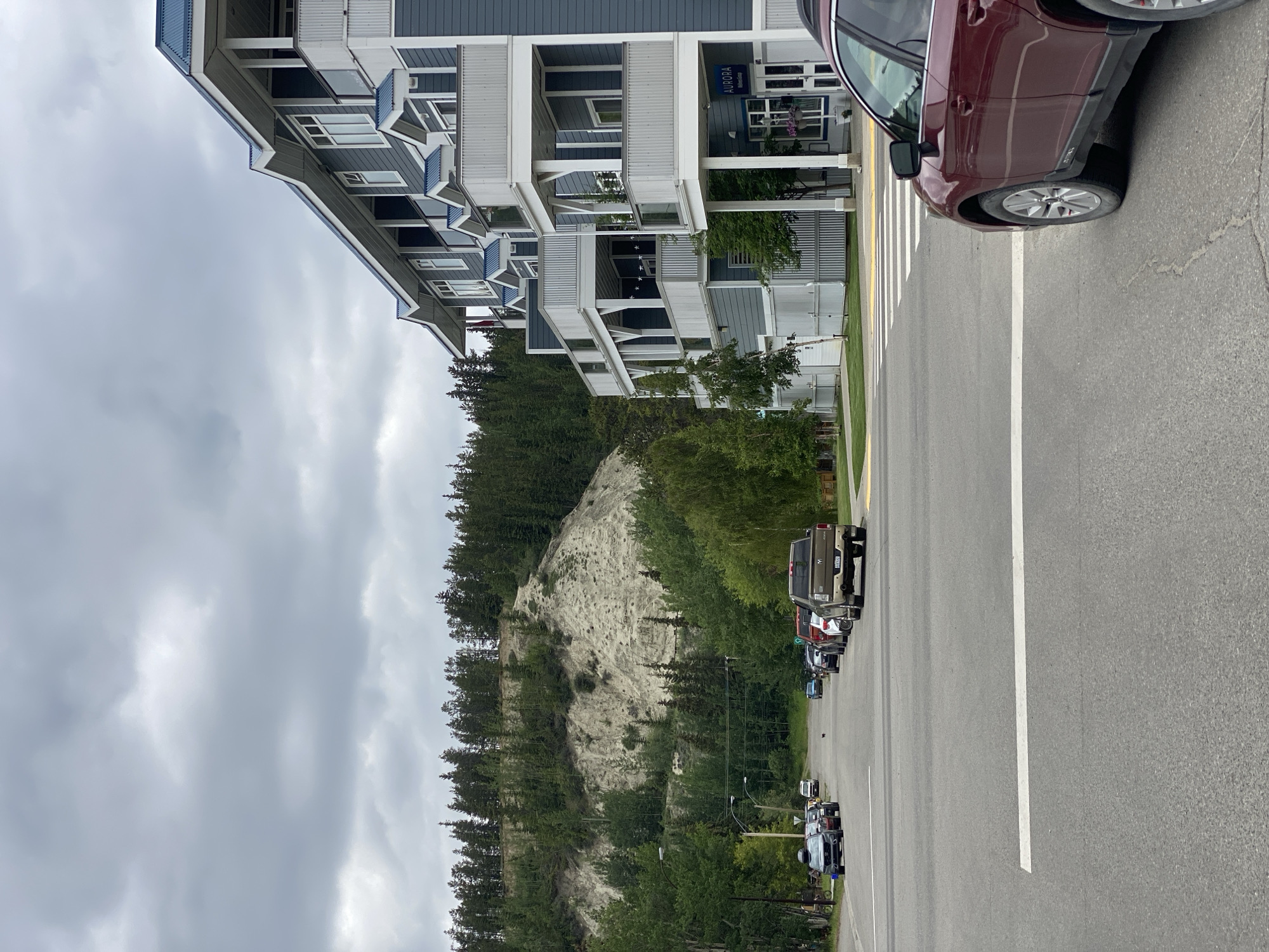 The image shows a street scene in Whitehorse, Canada, with a focus on a multi-story building on the right and a tree-covered hillside in the background. The building, with its dark blue siding and white trim, appears to be an apartment complex or condominium, featuring multiple balconies with white railings and blue roofs. A sign above the entrance reads "AURORA Wellness Group."
The street in the foreground is paved with asphalt and marked with white lines, indicating a crosswalk. A dark red SUV is parked in the foreground, partially obscuring the view of the crosswalk. On the left side of the street, several other vehicles are parked along the curb, including a silver SUV, a dark blue sedan, and a pickup truck.
The background features a steep, light-colored hillside covered with a dense forest of evergreen trees. The sky is overcast, with heavy gray clouds dominating the upper portion of the image. The overall impression is that of a residential area in a northern climate, possibly during the summer or shoulder season, given the foliage and the overcast sky. There are no people clearly visible in the image, and the focus is on the architecture, vehicles, and natural landscape.
