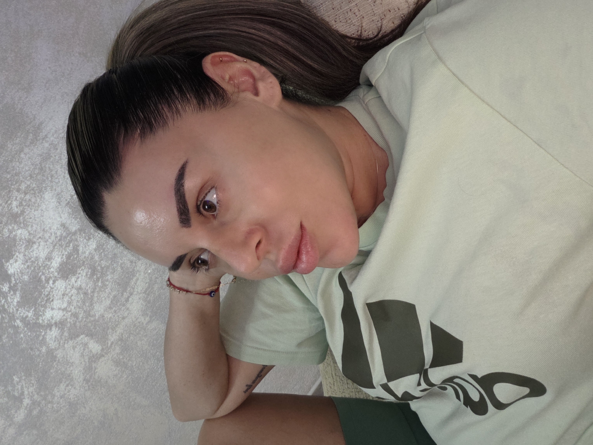 A medium close-up, slightly elevated shot shows a woman from the shoulders up, lying down and looking to her right. She has her left arm bent and resting behind her head. She is wearing a light green t-shirt with a dark grey graphic design. Her dark hair is pulled back into a ponytail. Her skin appears dewy, and she is wearing subtle makeup. A red bracelet with a blue evil eye charm is visible on her left wrist. The background is a textured, light grey wall. The scene is indoors, likely in a residential setting in the city of Peshtera, Bulgaria.