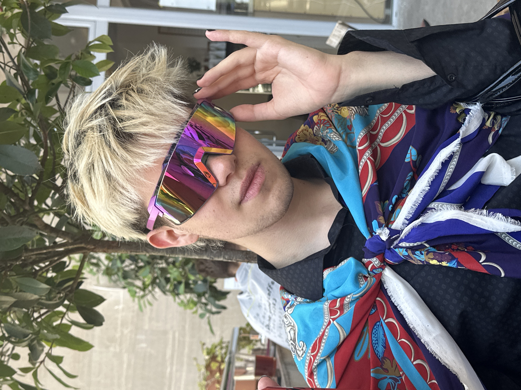 A young person with short, blond, spiky hair is captured in a close-up, horizontal shot. They are wearing large, angular, iridescent sunglasses that reflect a rainbow of colors. Their right hand is raised, with manicured black nails, and is gently touching the top of their head, near the sunglasses.
The person is wearing a black shirt with a subtle patterned texture and a brightly colored scarf tied around their neck and chest. The scarf features a bold, intricate design with elements of blue, red, white, and gold, including chain-like patterns and floral motifs. The overall aesthetic suggests a fashionable and perhaps avant-garde style.
The background is softly blurred, showing glimpses of greenery from a plant to the left and what appears to be a window or reflective surface behind. The lighting suggests it's daytime, possibly outdoors or in a well-lit indoor space with natural light. The person's expression is neutral, focusing attention on their accessories and style. There is no visible text in the image. The context of Budva, Montenegro is noted.
