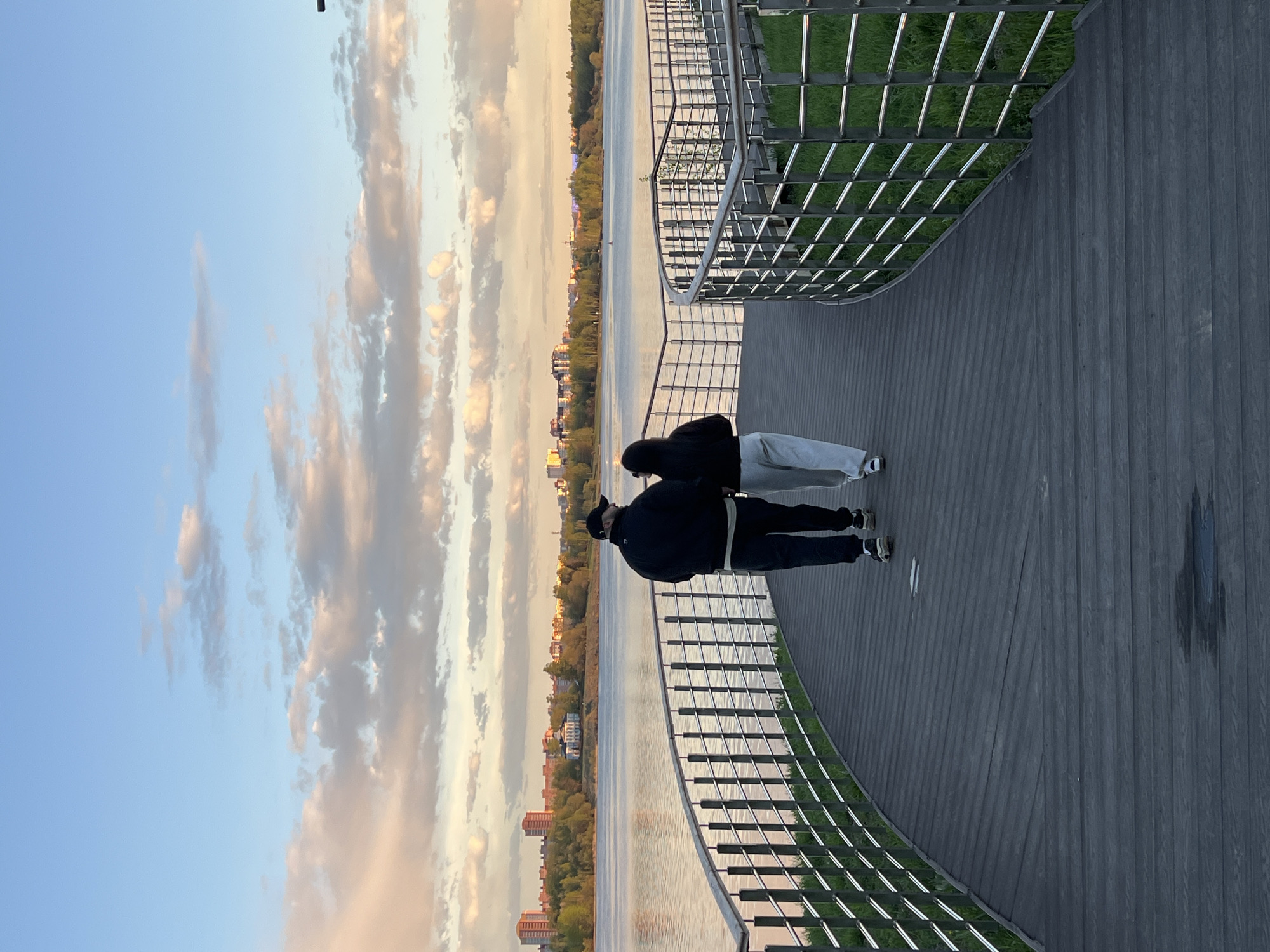 Two people stand on a wooden boardwalk next to a metal railing. Beyond the railing is water, a tree-lined shore, and buildings. The sky is blue with clouds. The scene is located in the city of Cherepovets, Russia.