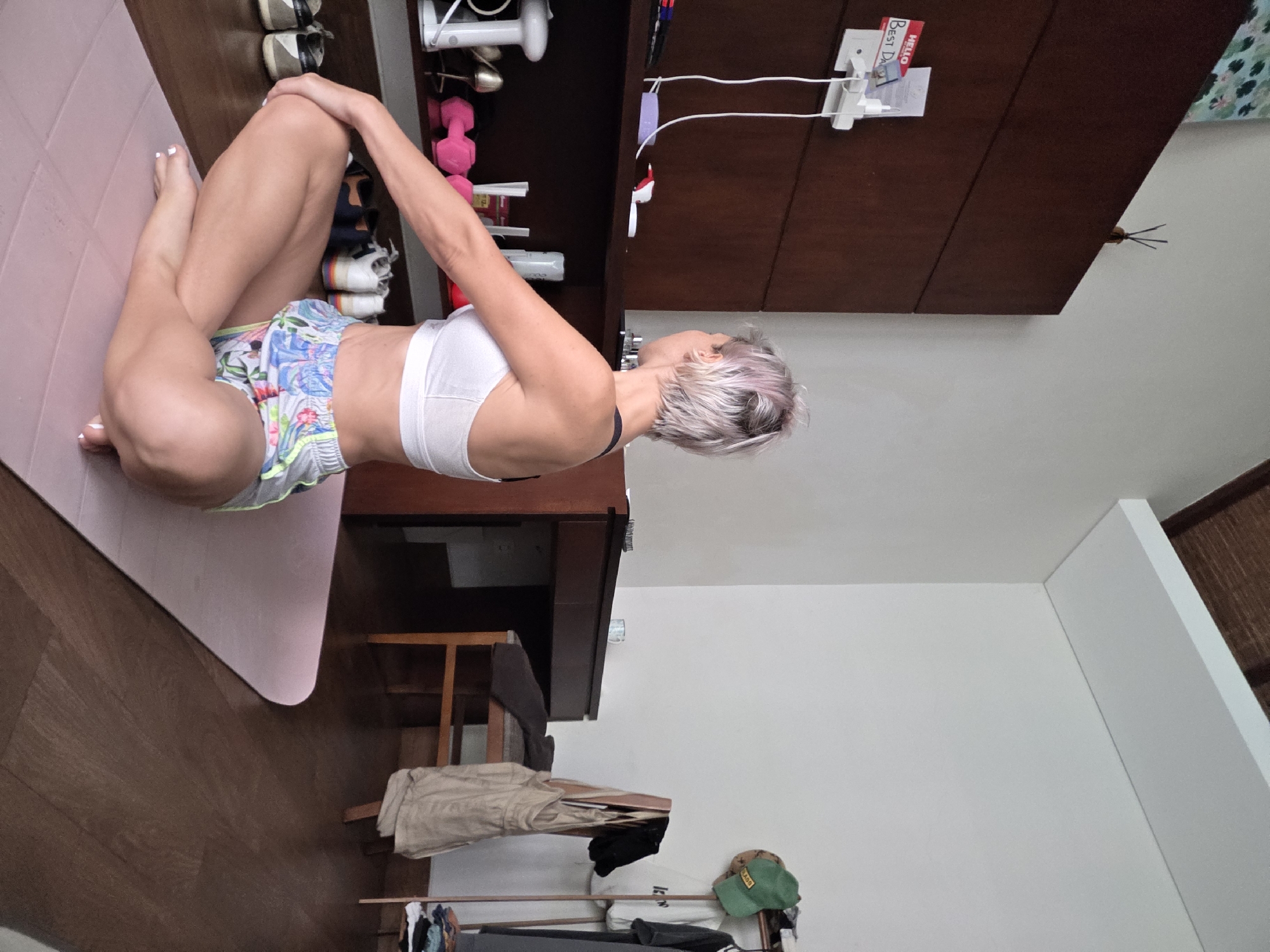 In a room with warm wooden floors, a woman with short, light-colored hair is performing a yoga pose on a pink mat. She is wearing a white sports bra and floral-patterned shorts. Her legs are crossed, and her hands are resting on her knees, with her back facing the camera.
The room appears to be a bedroom or a living space. To her left, a dark wooden shelf unit is visible, containing various items such as a white electronic device, pink dumbbells, and shoes. A desk with a chair is positioned behind her, and a clothes rack with garments and hats stands in the background. The walls are painted white, and a slatted wooden ceiling is visible towards the top of the frame. The overall impression is that of a private indoor setting, likely a hotel room or residence in North Kuta, Indonesia, given the humid climate suggested by the clothing and setting.