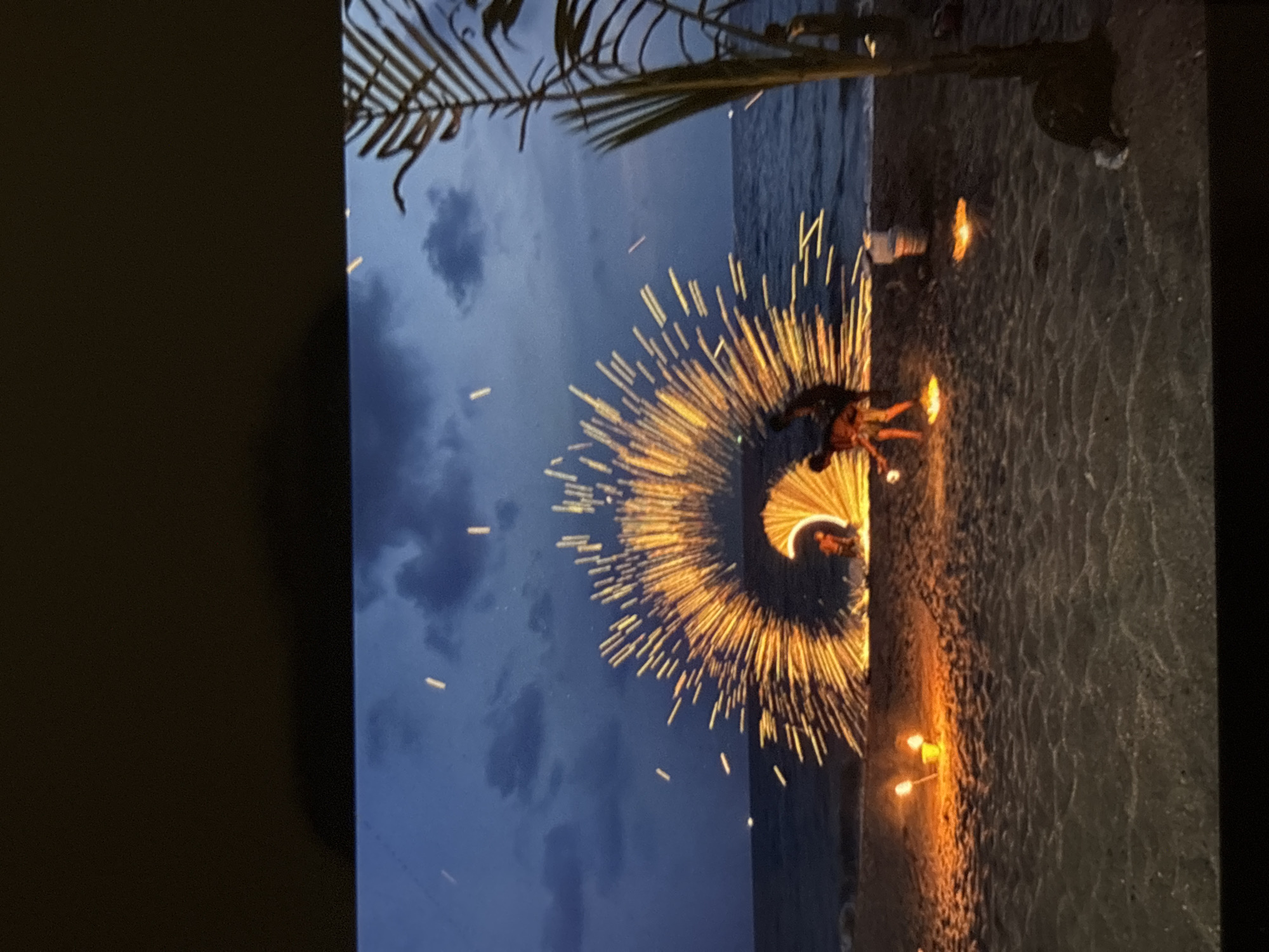 The image depicts a fire dancing performance on a beach in Chalong, Thailand, at dusk. The central focus is a performer creating a large, circular arc of sparks with a fire prop, which forms a bright, glowing halo against the darkening sky. Several other performers are visible, some engaging with fire, others standing. The beach is sandy, with the ocean and a faint horizon line in the background. Torches illuminate the sand in the foreground, casting an orange glow. A palm frond is visible on the right side of the frame, adding to the tropical setting. The sky is a deep blue, with scattered clouds and the faint lights of distant sparks. The overall scene is dynamic and captures the vibrant energy of the performance against a serene natural backdrop.