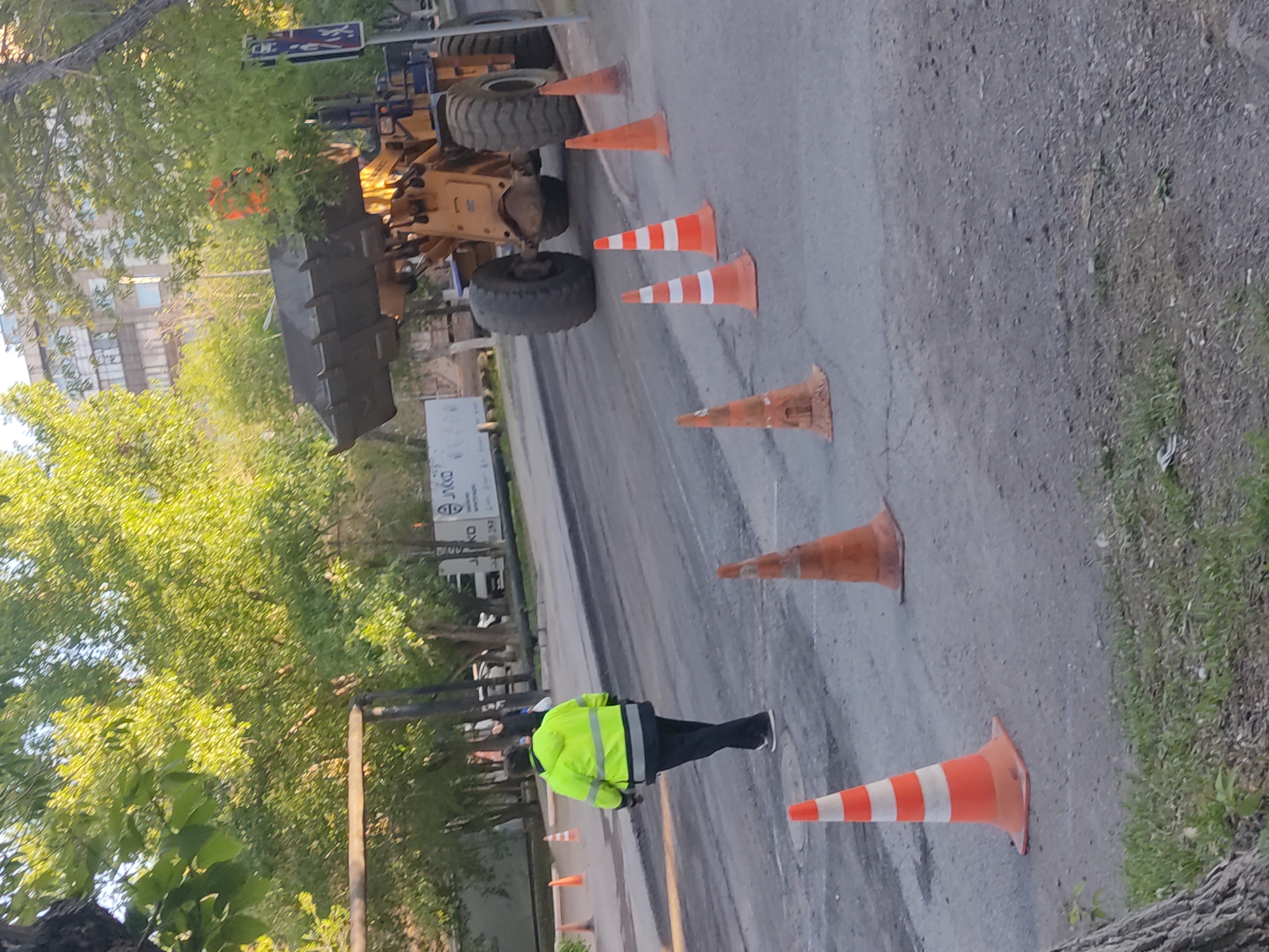 The image shows a street in Unknown, Unknown, possibly undergoing road work. A yellow construction vehicle is partially visible on the left side of the road, surrounded by several orange traffic cones. A person wearing a bright yellow safety vest is standing on the road near the cones. The road appears to be paved but with some visible imperfections and a gravelly shoulder. Trees are visible along the side of the road. A utility truck can also be seen in the background.