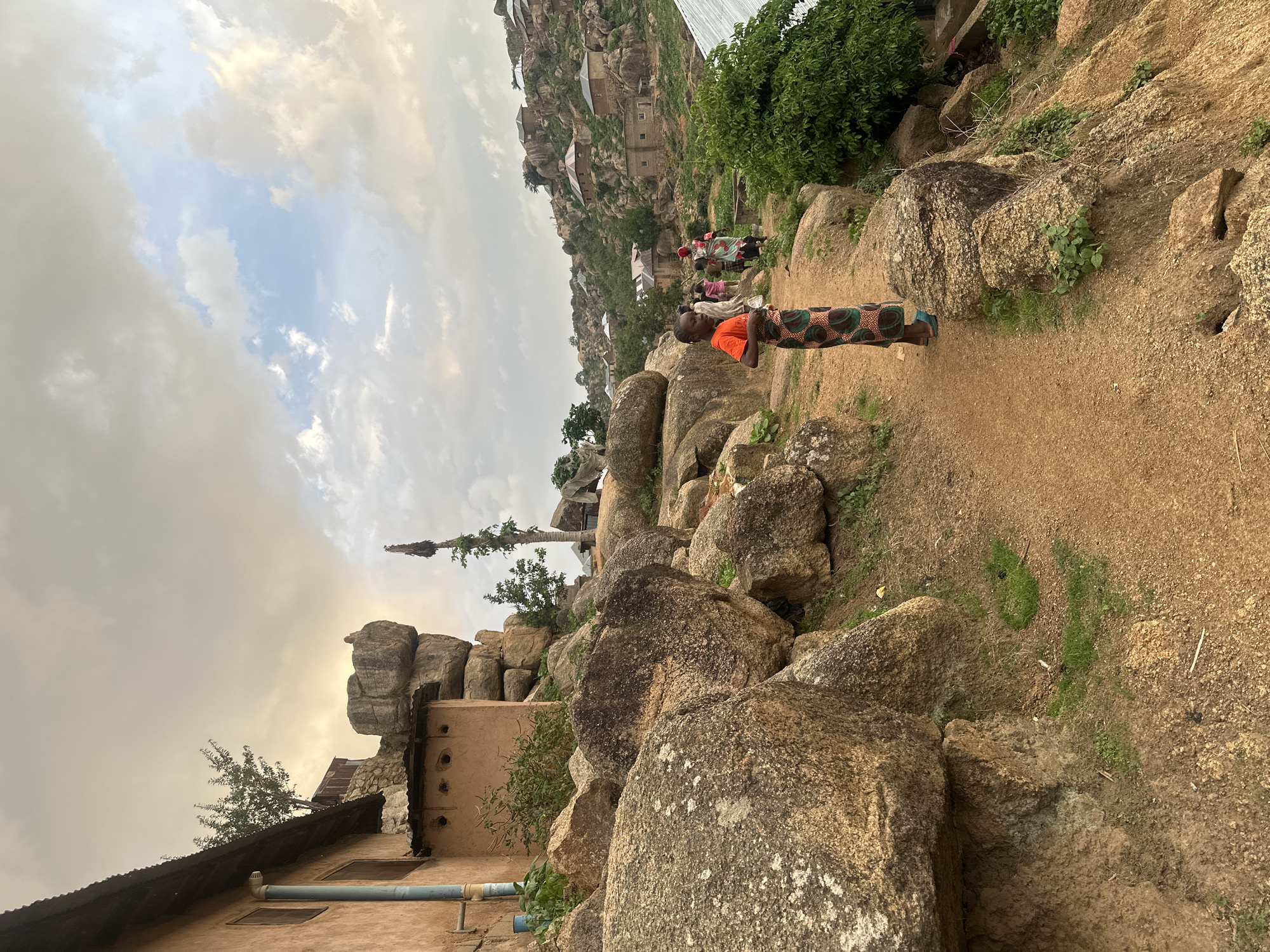 This image depicts a rural or semi-rural community nestled within a distinctive rocky, hilly landscape, characteristic of Jos, Nigeria. The scene is dominated by numerous large, weathered boulders and traditional dwellings built into or around these natural formations.
In the foreground, a person, likely a young woman or girl, stands on a reddish-brown dirt path. She is wearing an orange short-sleeved top and a patterned wrapper or skirt in green, brown, and black. Her stance suggests she is either walking or paused, with her body angled slightly away from the viewer but her head turned partially back.
The mid-ground and background reveal a scattering of houses, many featuring earth-toned walls and corrugated metal roofs. These structures appear integrated with the rocky terrain, some seemingly built directly against or atop the boulders. Sparse green vegetation, including bushes and small trees, grows between the rocks and around the houses. Further along the winding path and amidst the settlement, other indistinct figures can be seen, suggesting ongoing daily activities.
The sky is partly cloudy, with patches of soft blue visible through diffused white and gray clouds. The lighting is gentle and diffused, indicative of either a late afternoon or early morning, rather than harsh midday sun. No specific emotions are discernible from the distant figures. There is no visible text in the image. The overall impression is one of a community living in harmony with its unique, rugged natural environment.