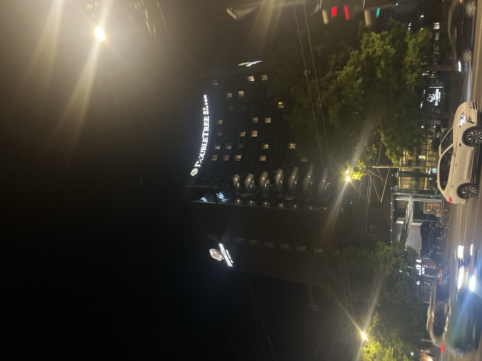 The image captures an urban scene at night in Yerevan, Armenia, featuring the DoubleTree by Hilton Yerevan City Centre hotel. The hotel's illuminated signage is visible on the building's facade. Street lights and vehicle headlights illuminate the scene. Cars are parked or moving along the street adjacent to the hotel. The presence of trees and the shapes of street lights create a blurred streetscape along with signs. The overall impression is that of a moderately busy city scene at night.