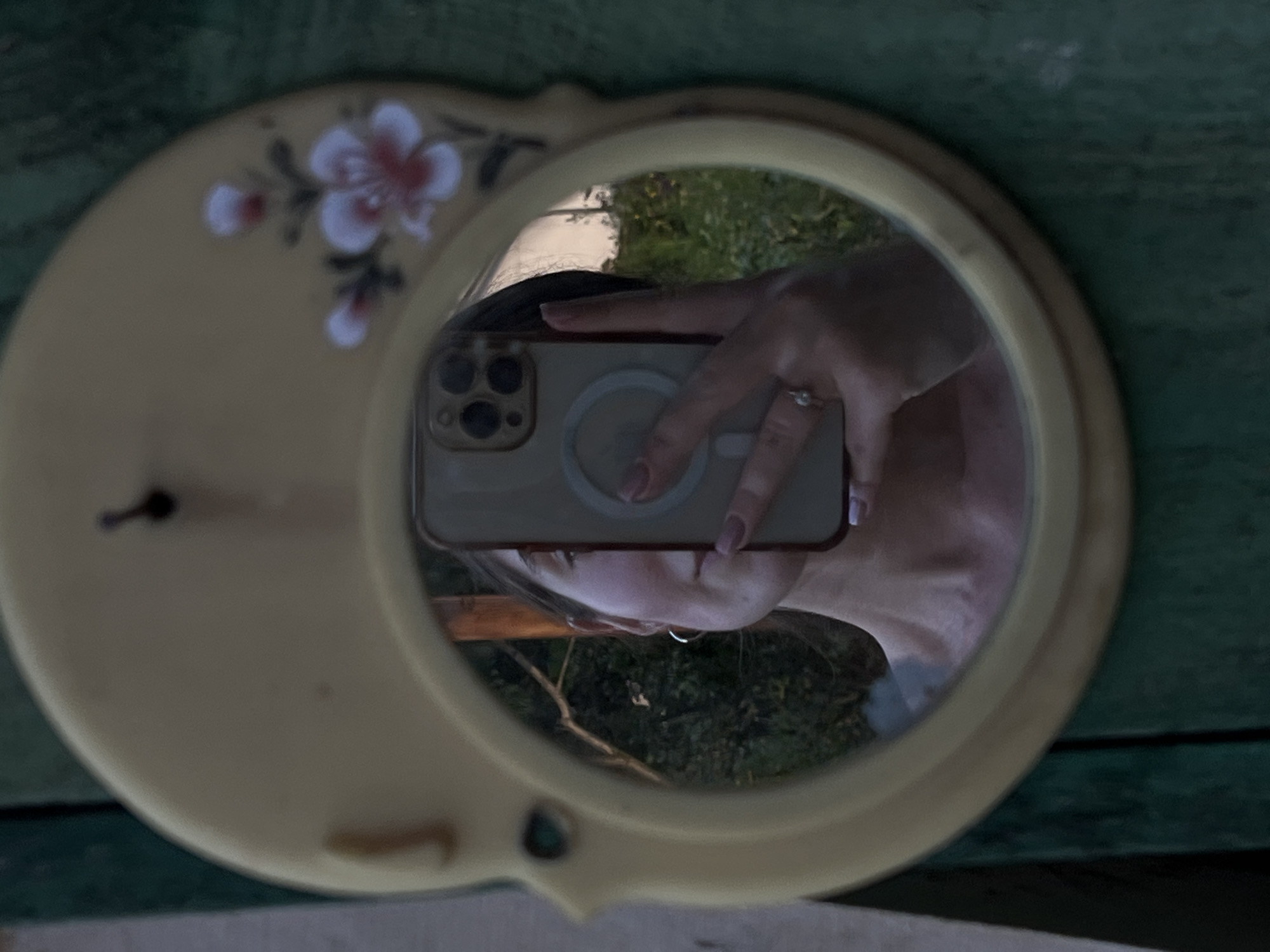 The image shows a young woman taking a selfie in a round mirror. The mirror is oval-shaped and has a yellow frame with a floral design. The woman is holding a smartphone in her hand, and her reflection is visible in the mirror. She is wearing a ring on her finger and has hoop earrings. The background appears to be outdoors, with greenery visible in the reflection. The mirror is hanging on a green, textured surface, possibly a wooden wall. The overall impression is of a casual, outdoor setting, possibly a garden or yard. There is no information in the image to suggest the location is the city of Dys, Poland.