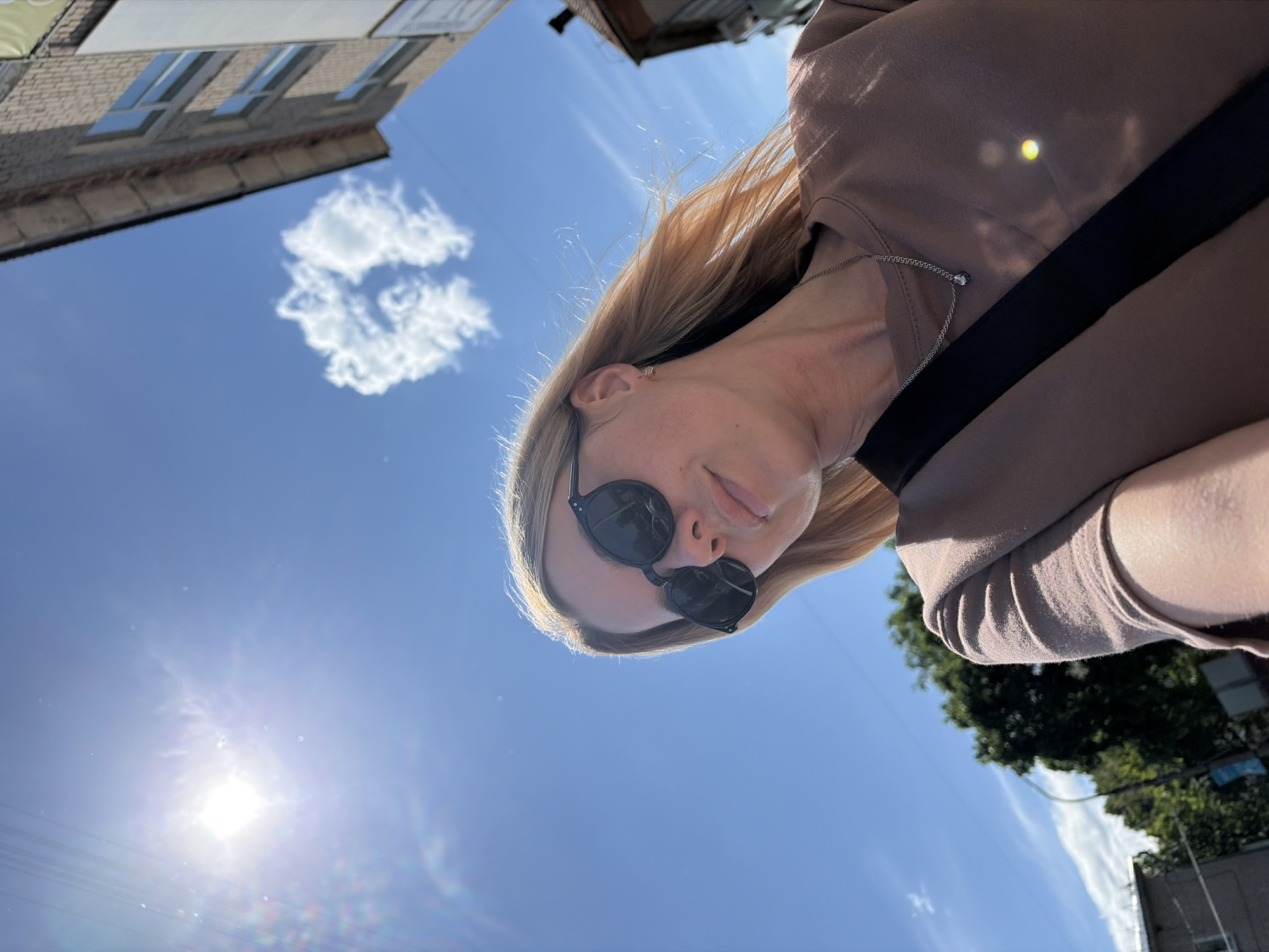 The image captures a sunlit outdoor scene in the city of Chaikovsky, Russia, featuring a woman from a low-angle perspective. The upper two-thirds of the frame are dominated by a clear, bright blue sky with scattered white clouds. The sun is prominently visible in the upper left, emitting a strong lens flare and distinct light rays. A singularly shaped, amorphous white cloud is located in the upper right quadrant.
A woman with blonde hair, styled to appear wind-swept, occupies the lower portion of the image, angled slightly upwards and towards the viewer's left. She wears dark, round-framed sunglasses that obscure her eyes, and her lips are closed in a subtle smile. Her attire consists of a plain, short-sleeved, light brown t-shirt. A thin silver chain necklace with a small, spherical pendant rests on her collarbone. A black strap, consistent with a bag strap, diagonally crosses her chest from her left shoulder downwards.
In the upper right background, a portion of a multi-story building constructed of light brown brick with visible window frames is present. To the left and in the mid-ground, dense green foliage from trees is visible, with a streetlight pole partially obscured amongst them. Faint structures are also discernible in the distant lower-left background. The overall lighting indicates a bright, sunny day.