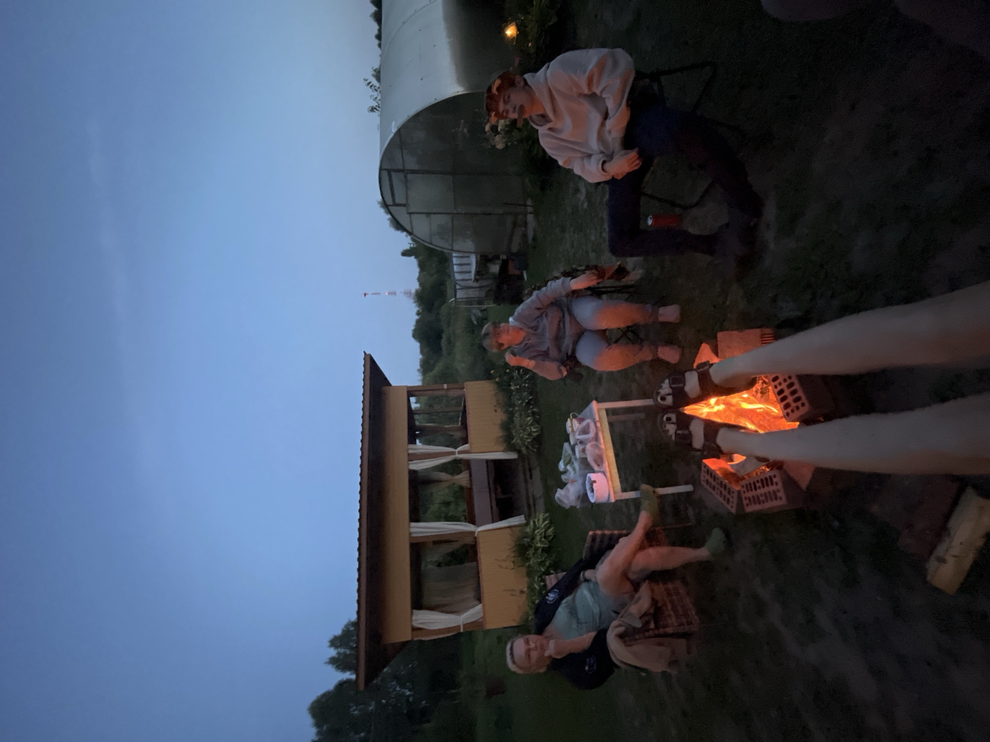 In Суроватиха, Russia, at dusk, four individuals are gathered around a campfire in a backyard setting. The scene is illuminated by the warm glow of the fire, and the sky is a deep twilight blue.
To the left, a woman with blonde hair is seated in a wicker chair, wearing a light blue top and dark jacket. She is looking to her left, with her hand on her chin. In front of her is a small table with various items, including what appears to be food and drinks.
In the center, another woman is seated in a chair, wearing a grey hooded sweatshirt and blue pants. She is looking forward.
To the right, a young man with reddish hair is seated in a chair, wearing a light grey hooded sweatshirt and dark pants. He is holding a cigarette or similar object to his mouth and looking to his left. A red can is visible on the ground near his chair.
In the foreground, two sets of legs are visible, wearing sandals, extending towards the campfire. The campfire is built with bricks and is actively burning.
In the background, a light-colored gazebo with white curtains is situated. Further back, a greenhouse structure is visible, and beyond that, trees and a distant tower with red lights. The ground is covered in grass and dirt.