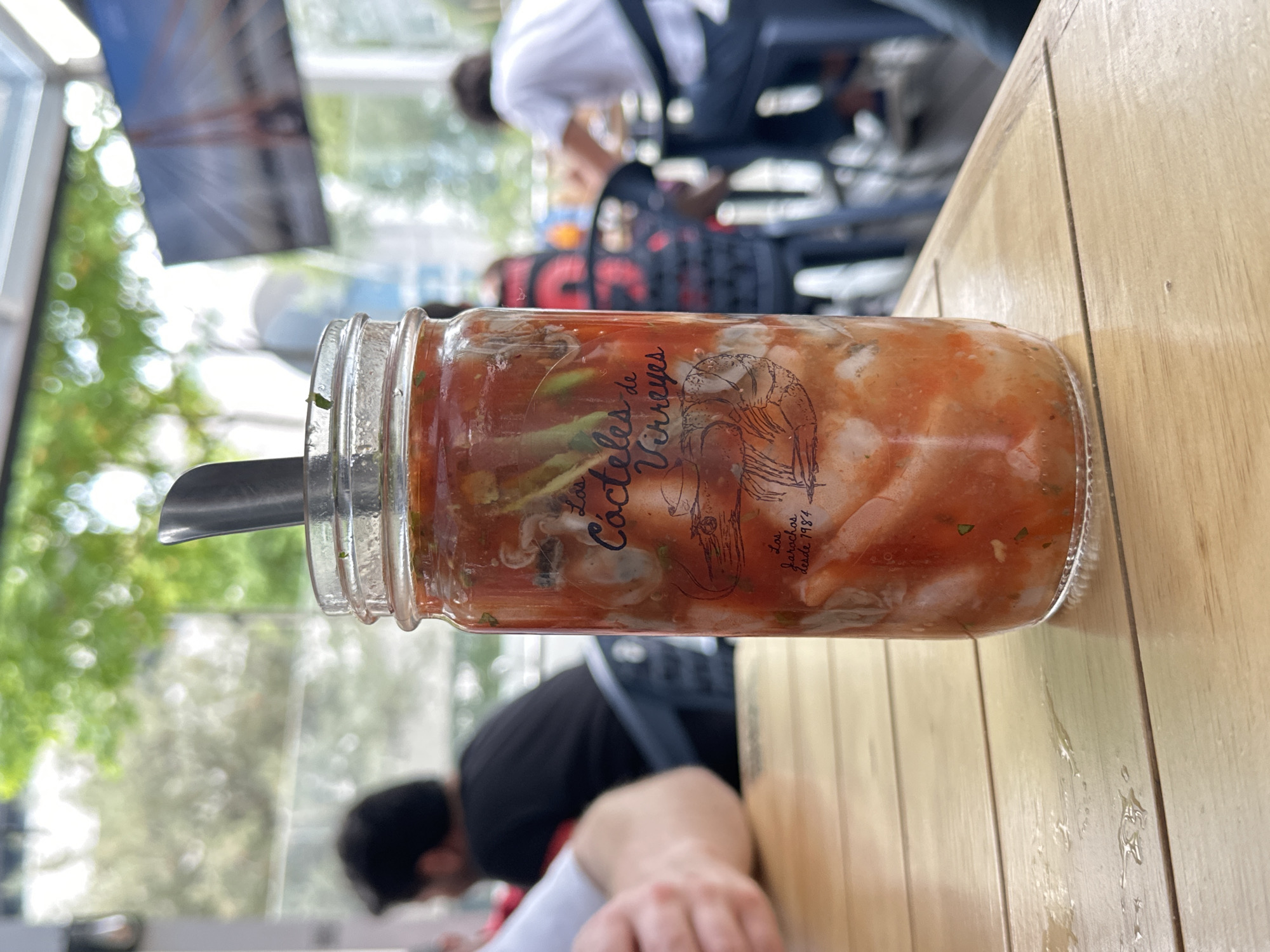 A glass jar filled with a red, viscous substance, possibly a cocktail, rests on a light-brown wooden table. A silver spoon is inserted into the jar. The jar is labeled "Los Cacteles de Virreyes," and underneath, there is the line "Los jarachos desde 1984". In the background, people are seated at tables in an outdoor seating area. Foliage and architectural elements are visible in the background, suggesting an urban environment, specifically, a restaurant in Mexico City, Mexico.