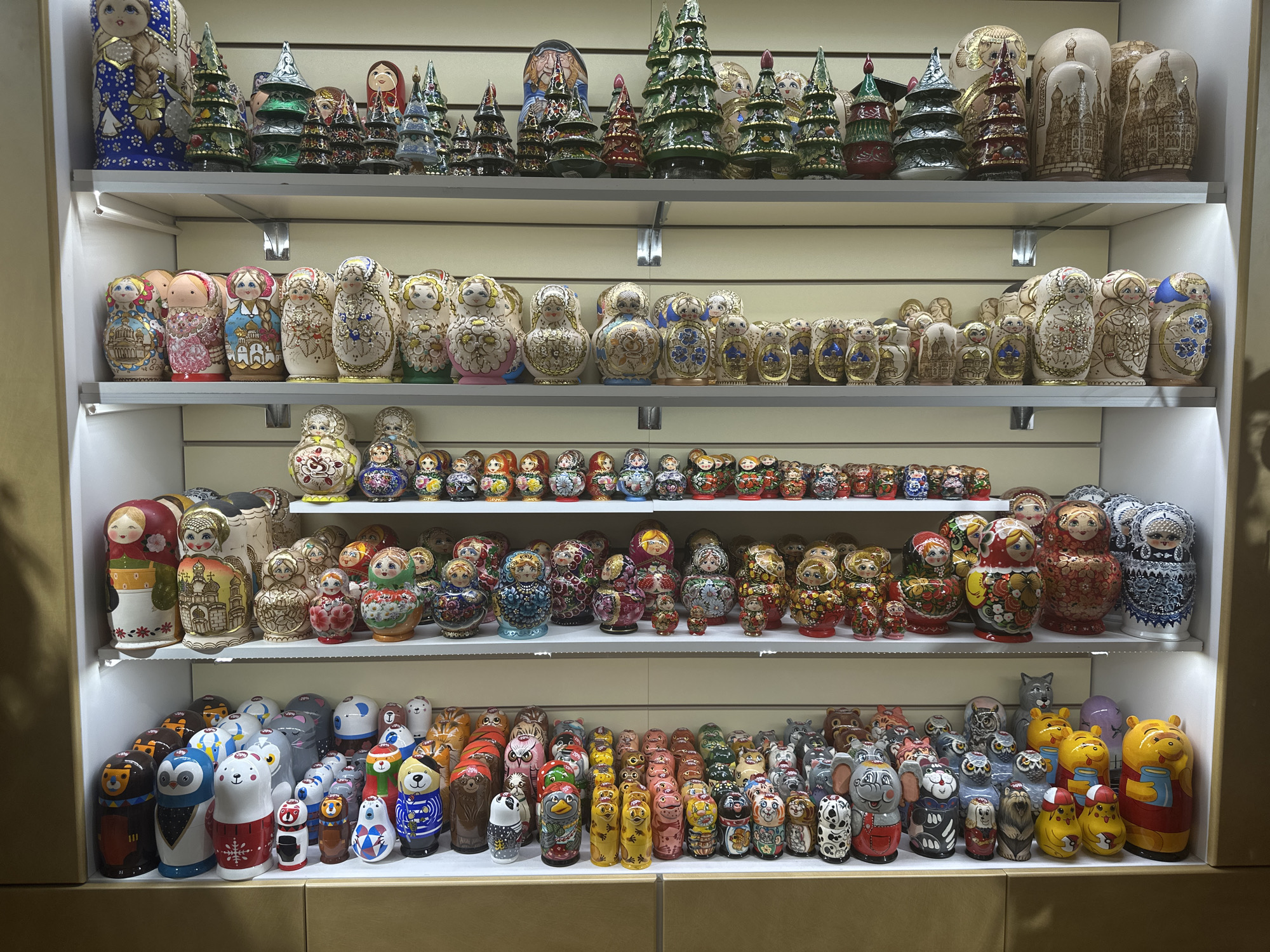 The image shows four shelves full of nesting dolls (Matryoshka dolls) and other decorative objects. The top shelf features nesting dolls alongside miniature Christmas trees. The second shelf contains a wide array of nesting dolls with varied designs and colors. The third shelf is filled with smaller nesting dolls. The bottom shelf displays nesting dolls painted to resemble various animals and characters. The scene suggests a shop or market stall in Saint Petersburg, Russia, where Matryoshka dolls are popular souvenirs. The environment is well-lit, highlighting the colors and details of the objects. There are no people visible in the image.