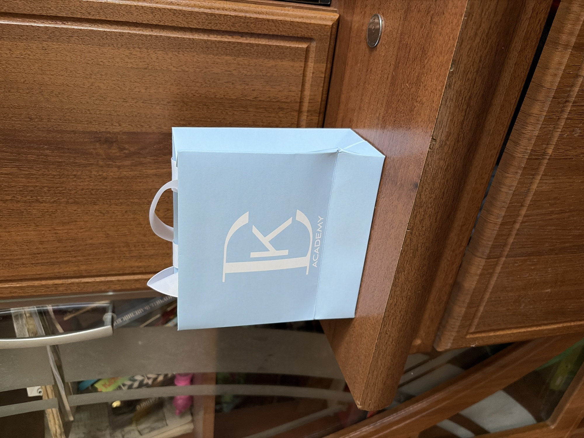 A light blue paper shopping bag with white ribbon handles sits on a wooden surface. The bag features a large white 'D' logo with a 'K' inside it, and the word "ACADEMY" printed below. To the left of the bag is a glass-fronted cabinet filled with books and other items. To the right of the bag is a wooden cabinet with carved details. A coin rests on the wooden surface in front of the cabinet. The scene appears to be indoors, possibly in a room with wooden furniture.