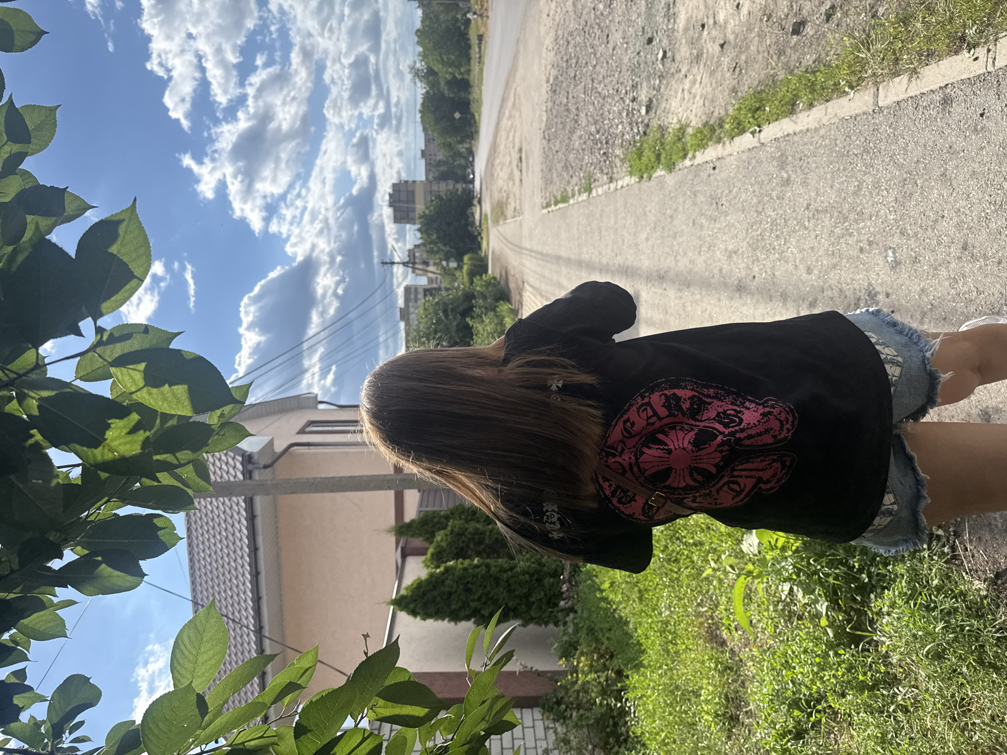 A person with long brown hair wearing a black t-shirt with a large pink graphic on the back and denim shorts walks along a sidewalk in Dnipro, Ukraine. The sidewalk is next to a road with a gravel shoulder. In the background, there are buildings, trees, and a cloudy blue sky. Green foliage from a tree hangs over the scene from the left side of the frame.