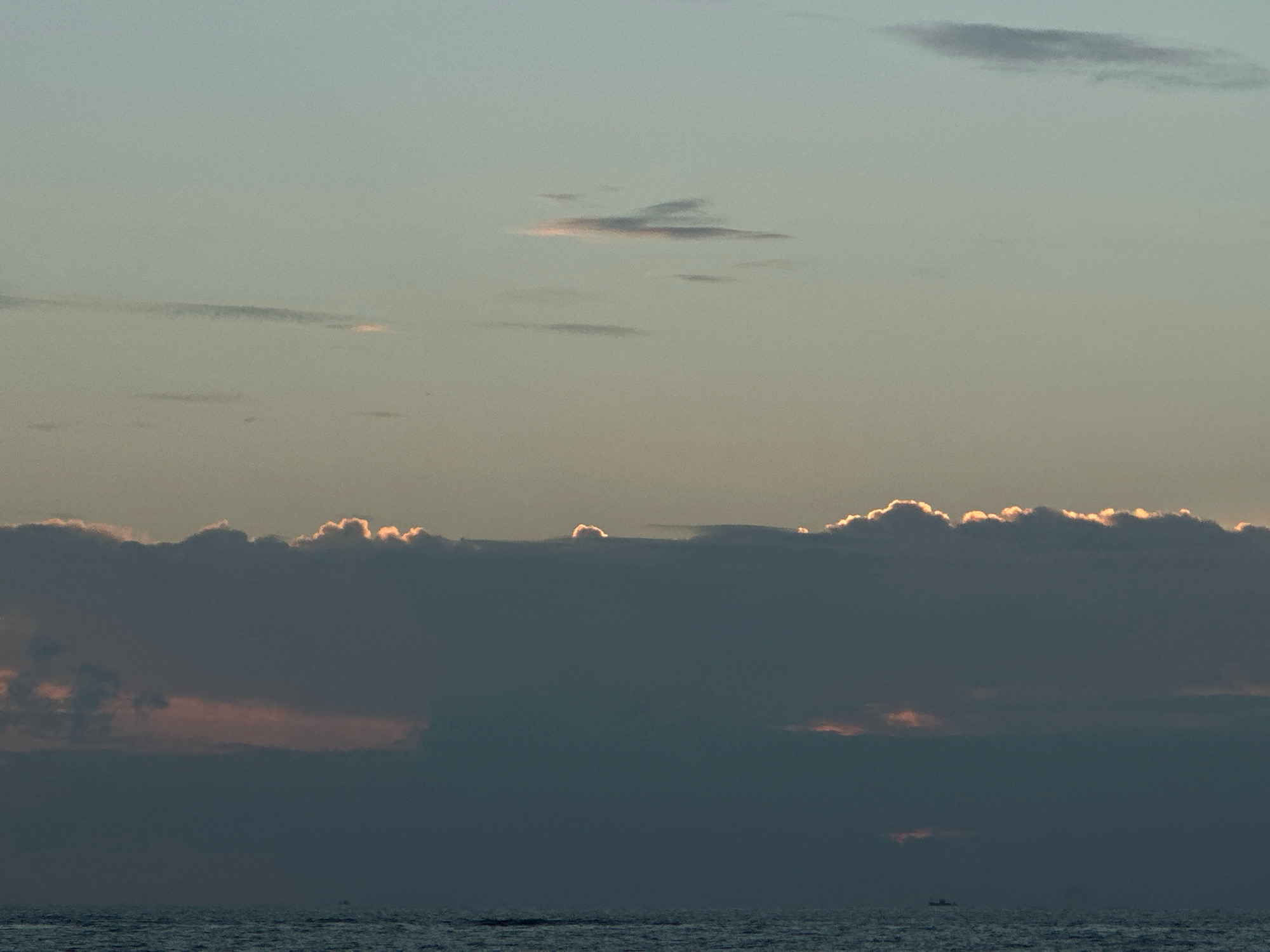 The image shows a serene seascape at dusk, possibly from the coast of Arambol, India. The sky is a gradient of soft blues and grays, with wispy clouds scattered across it. The sun, low on the horizon, casts a warm, golden glow on the edges of some of the clouds, creating a beautiful contrast with the darker, more substantial cloud bank that dominates the middle of the frame. This lower cloud formation appears heavy and almost imposing, obscuring the horizon. Below the clouds, the ocean stretches out, its surface gently rippling with the subtle ebb and flow of waves. The water is a deep, dark blue, reflecting the muted tones of the sky. In the distance, a solitary boat is visible, a small silhouette against the vast expanse of water, adding a sense of scale and tranquility to the scene. The overall impression is one of peacefulness and natural beauty.