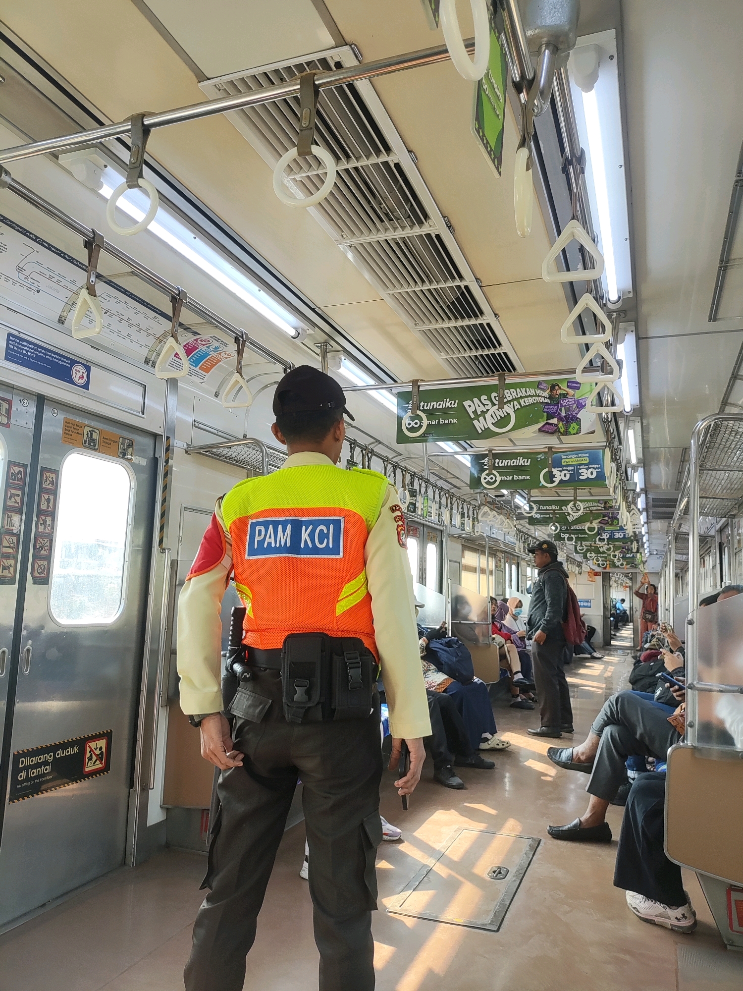 A man in an orange and yellow vest labeled "PAM KCI" stands facing away from the camera, inside a train car. He wears a black cap, dark pants, and a utility belt. The train car is populated with passengers, some seated, others standing. Ads are visible on the ceiling of the car. The floor is a light brown color, and the walls are metallic. There is a door to the left with a sign that reads "DILARANG DUDUK di lantai" (No sitting on the floor). The overall scene depicts a moment within public transportation. This scene is located inside a train in the city of Bekasi, Indonesia.