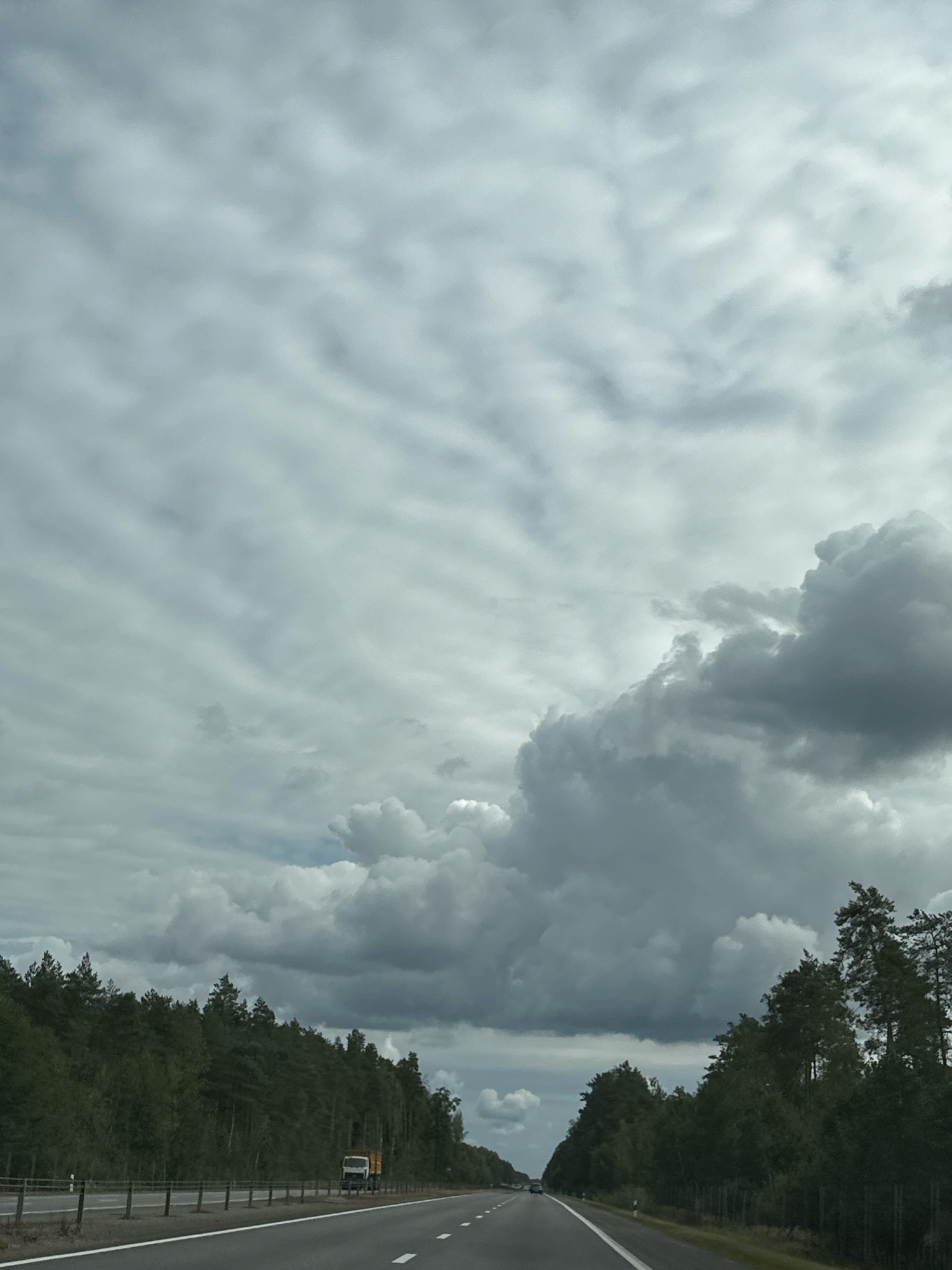 The image depicts a highway scene under a cloudy sky. The road is paved and appears to be multi-lane. On the left side of the road, there is a dense forest of evergreen trees. On the right side of the road, the forest continues. A white truck with a yellow cargo area is parked on the shoulder of the road on the right. The sky is filled with numerous clouds, varying in shade from light gray to dark gray, suggesting an overcast or possibly stormy weather condition. The overall scene is of a rural or semi-rural highway. The location provided is Вяззеўскі сельскі Савет, Belarus.