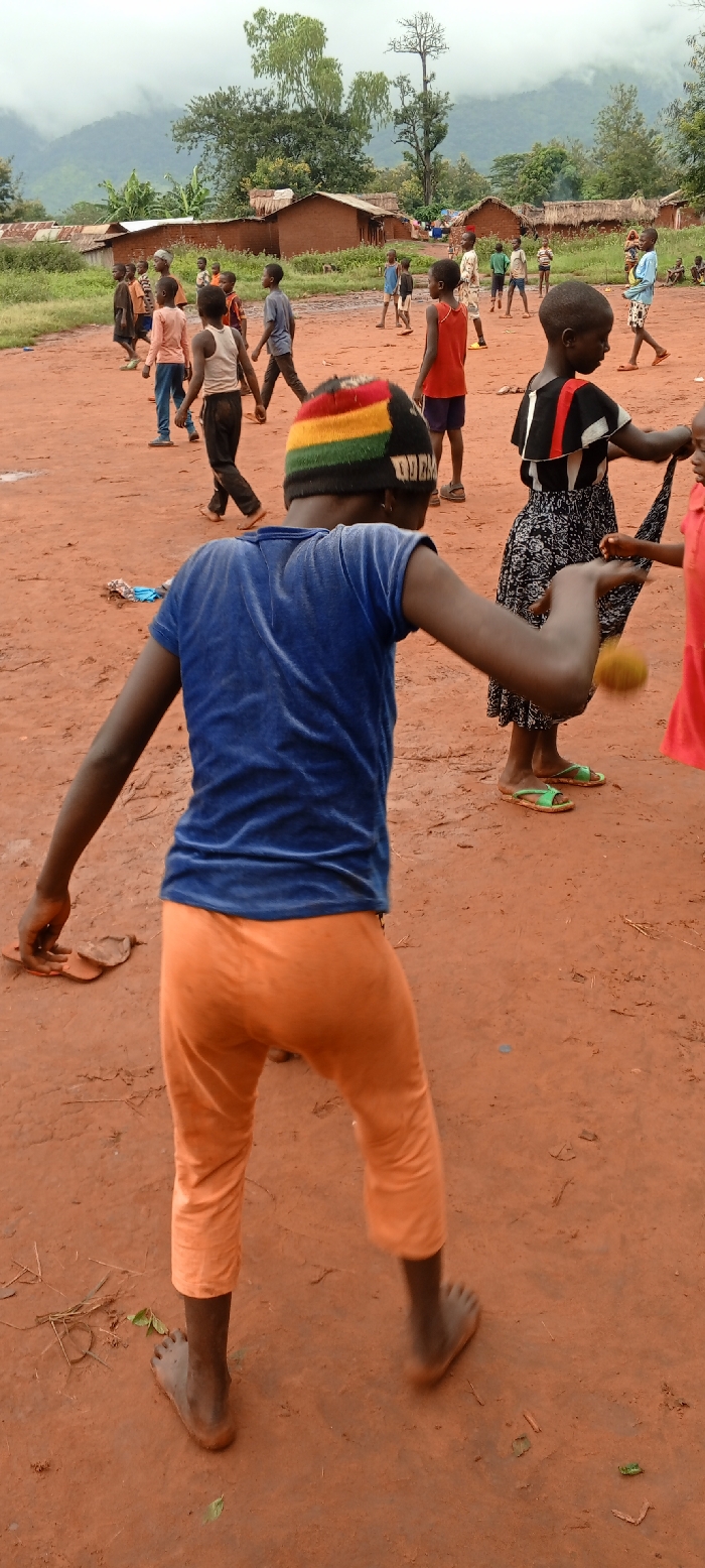 An outdoor scene in Ngalula, Democratic Republic of the Congo, depicts approximately fifteen children gathered on a reddish-brown dirt field under an overcast sky. In the foreground, a child, viewed from behind, wears a blue velvet short-sleeved top, orange cropped pants, and a black knit beanie with horizontal red, yellow, and green stripes and white text. This child is barefoot, with arms slightly extended and bent, suggesting motion. To the right, another child in a black and white patterned top and skirt wears green flip-flops and interacts with a smaller child in a red shirt, appearing to exchange a small, light-brown, spherical object.
In the midground and background, numerous other children, ranging in age, are dispersed across the expansive dirt area. Their attire varies, including t-shirts, shorts, and dresses, with some children wearing sandals and others barefoot. Behind the children, several single-story structures constructed from reddish-brown mud bricks with corrugated metal or thatched roofs line the edge of the dirt field. Beyond the structures, dense green foliage and tall trees are visible, preceding large, mist-covered mountains under a diffused, gray sky in the far background. The ground exhibits minor irregularities and scattered natural debris such as small leaves and twigs.